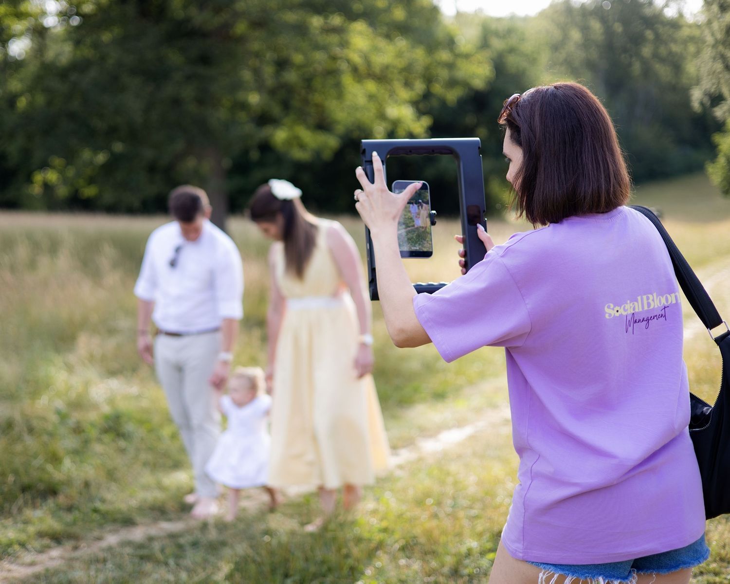 Woman in purple shirt photographing family with tablet in sunny green meadow. Social Bloom Management, South London