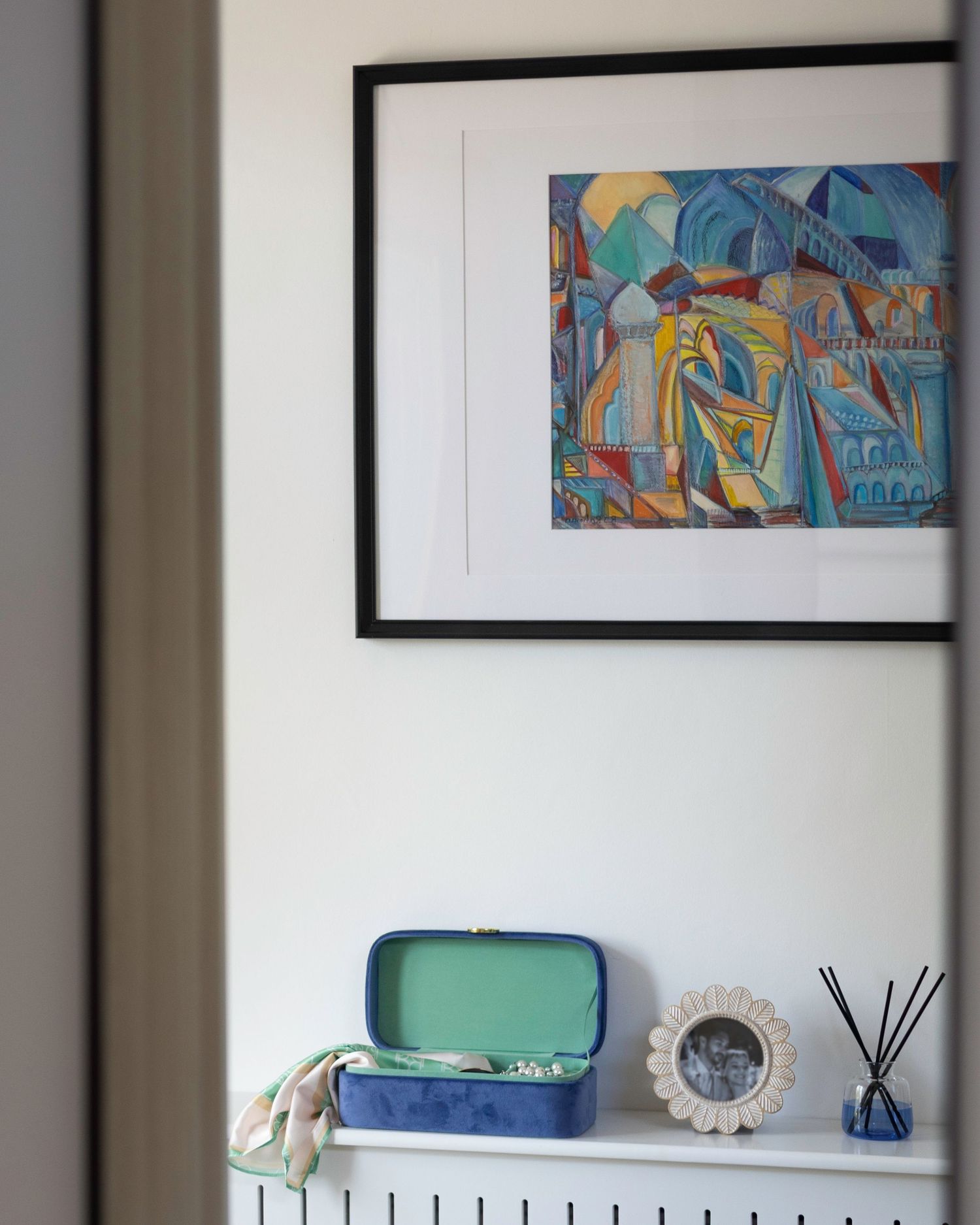 Colorful abstract framed artwork above a white shelf with a teal jewelry box, photo frame, and reed diffuser. Brand Photography South London