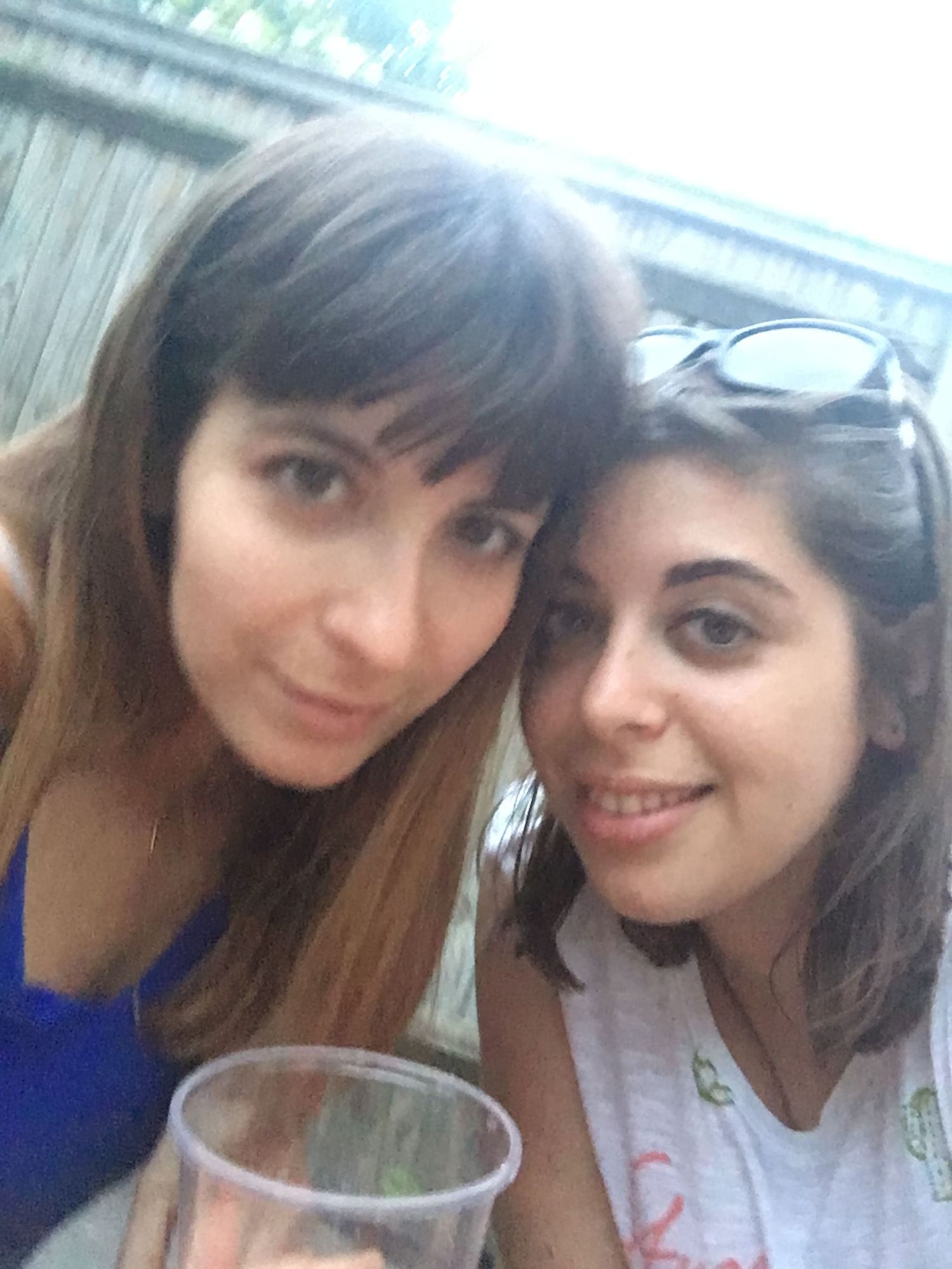 Two young women smiling together outdoors at a casual gathering, one holding a clear plastic cup.