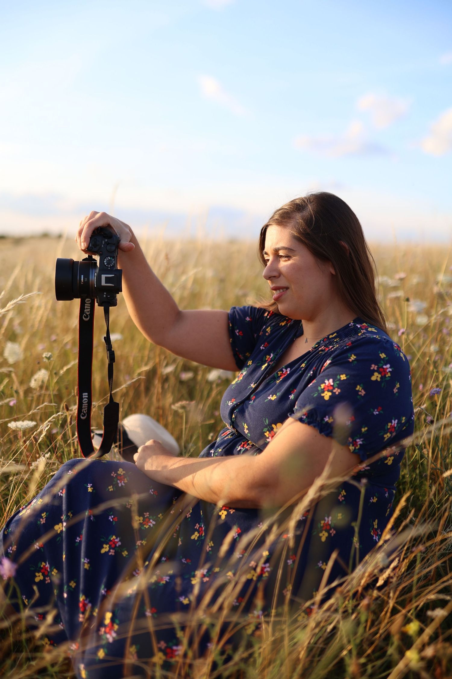 Young woman in floral blue dress sitting in golden field holding Canon camera on tripod. South London