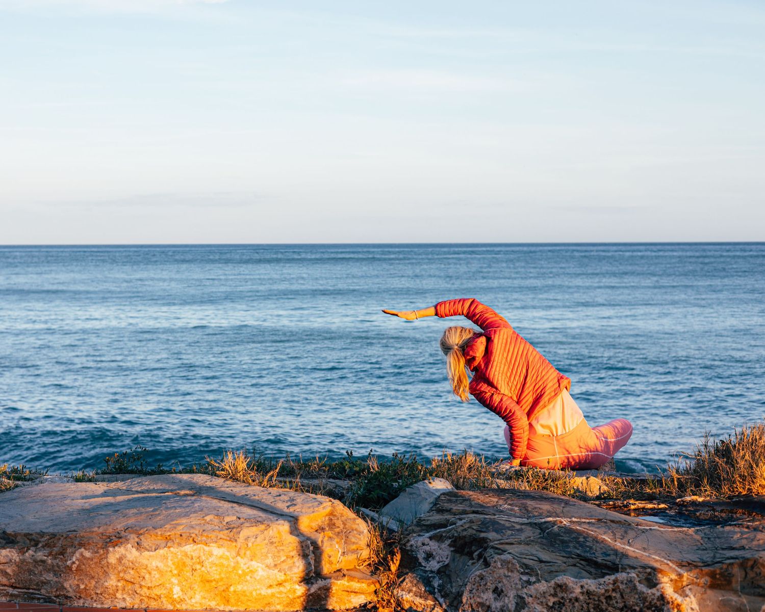 Person in orange outfit practicing yoga on rocky coastal cliffs with ocean horizon in background. Yoga by Ocean, Imperia, Italia