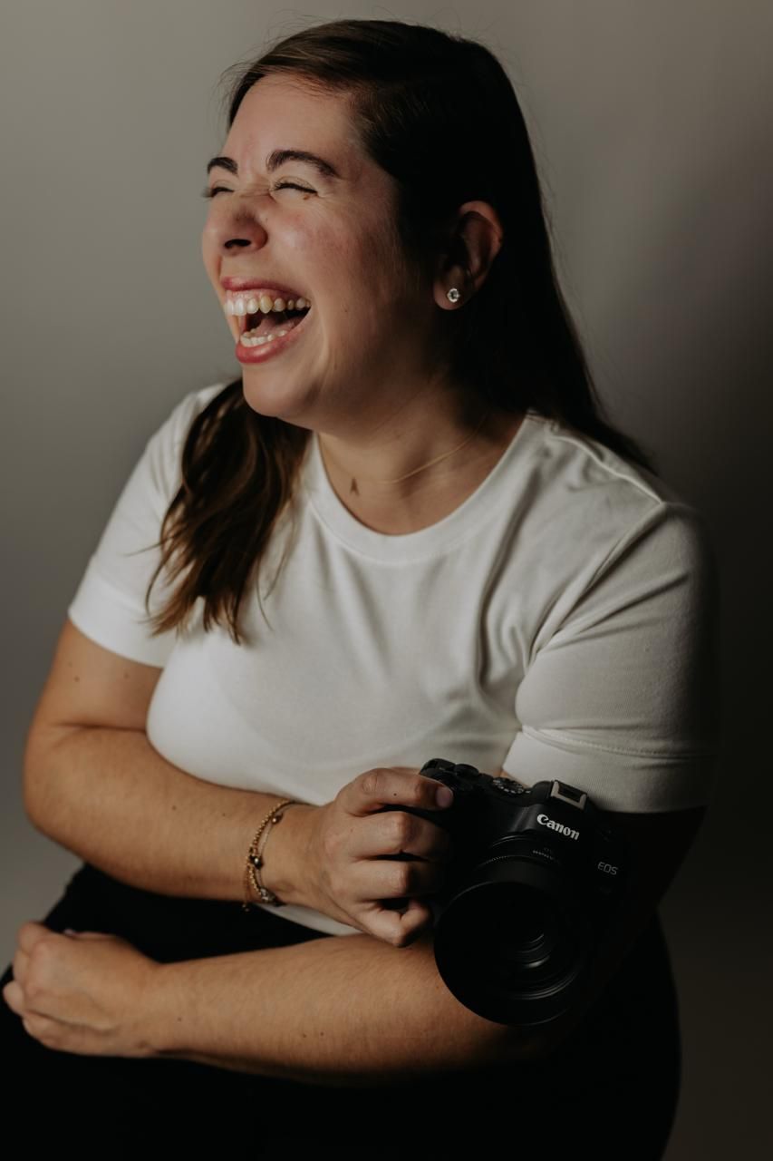 Laughing brunette woman holding Canon camera wearing white t-shirt against gray studio background. South London