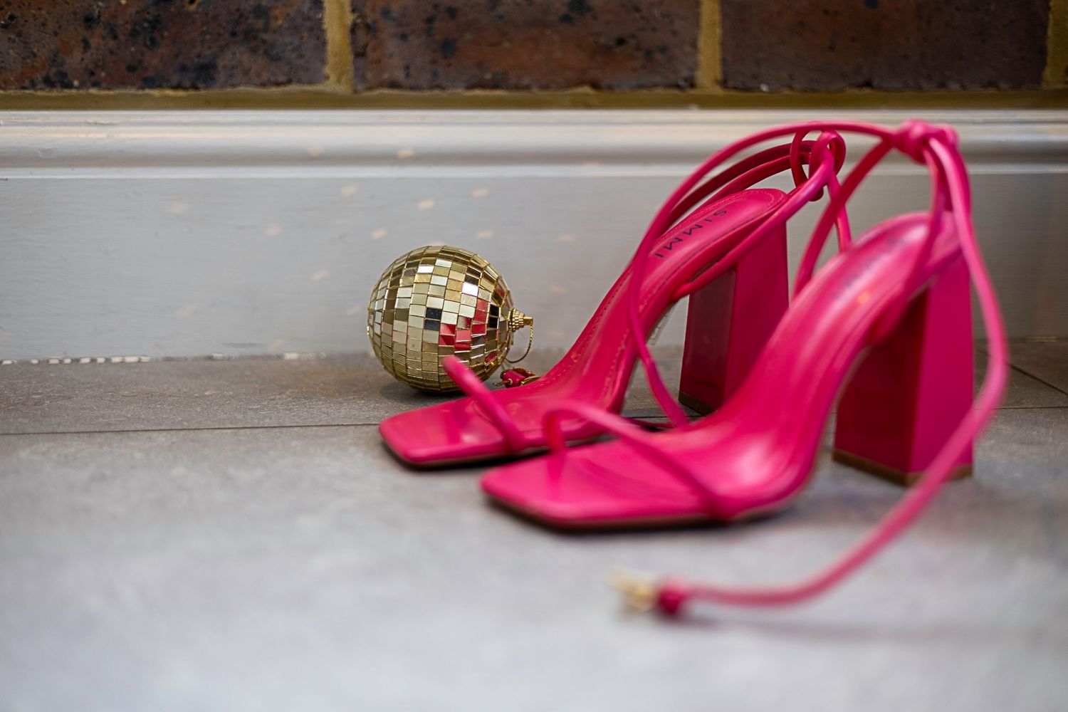 Hot pink strappy block heel sandals next to a small gold disco ball on white surface.