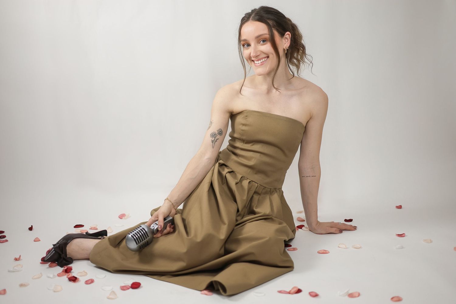 Smiling brunette woman in strapless olive dress sitting among red rose petals holding microphone. Brand Photography South London