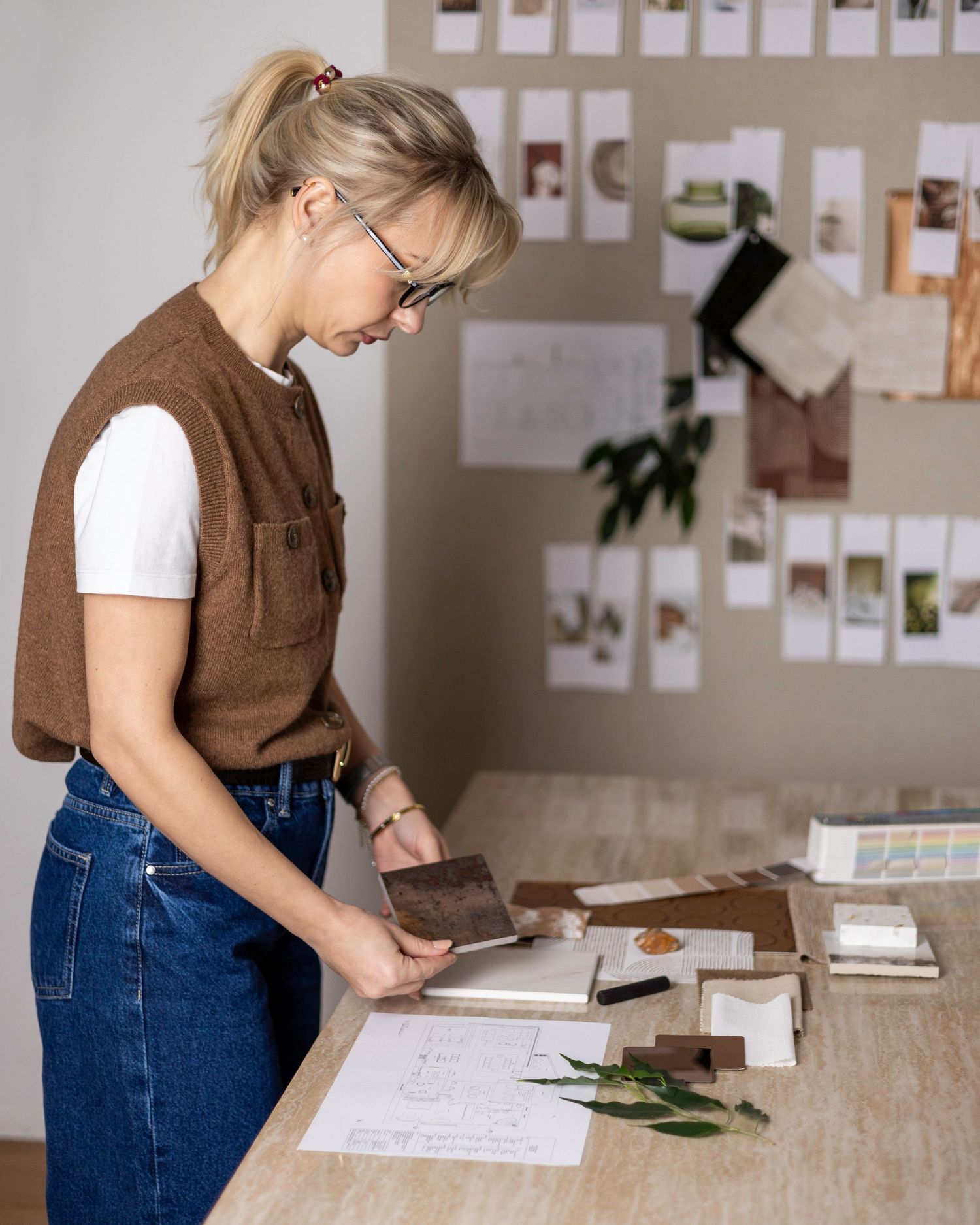 Blonde woman in brown vest reviewing design samples at wooden desk. South London