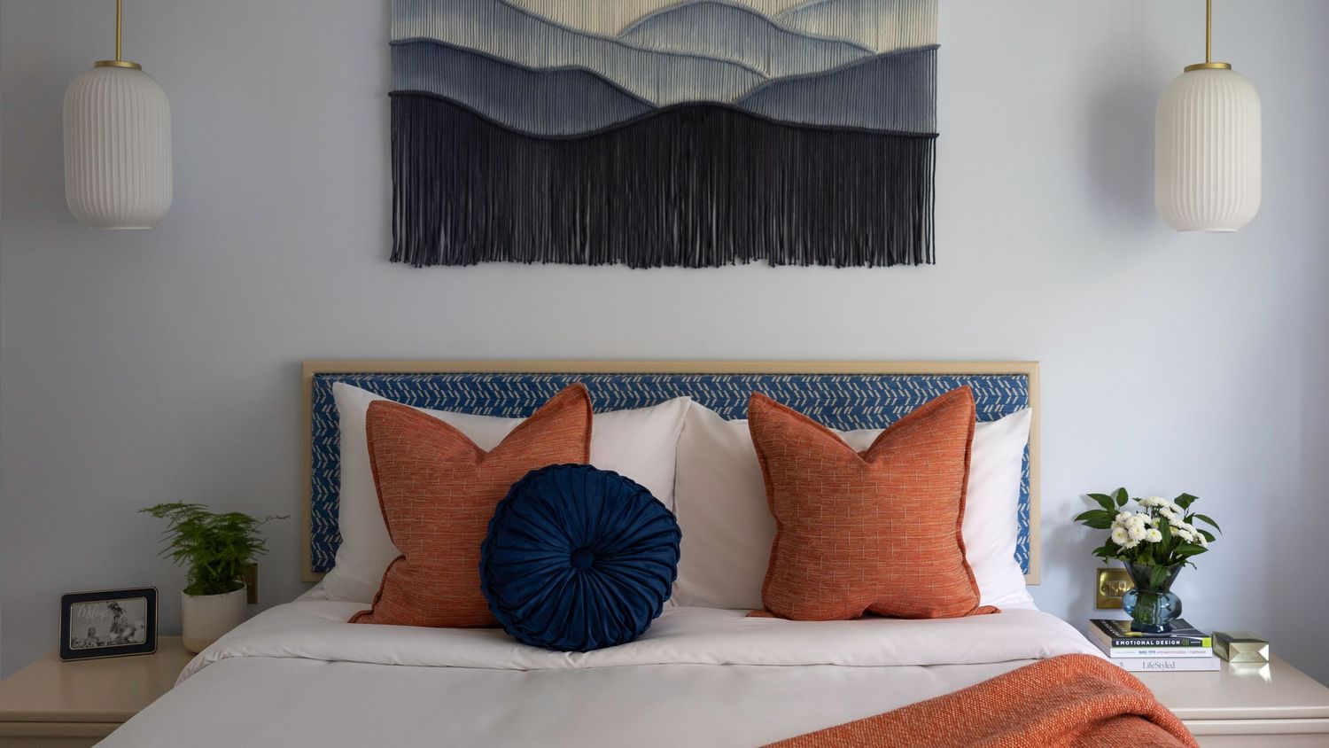 Stylish bedroom with blue upholstered headboard, orange and navy pillows, and macrame wall art. Brand Photography South London