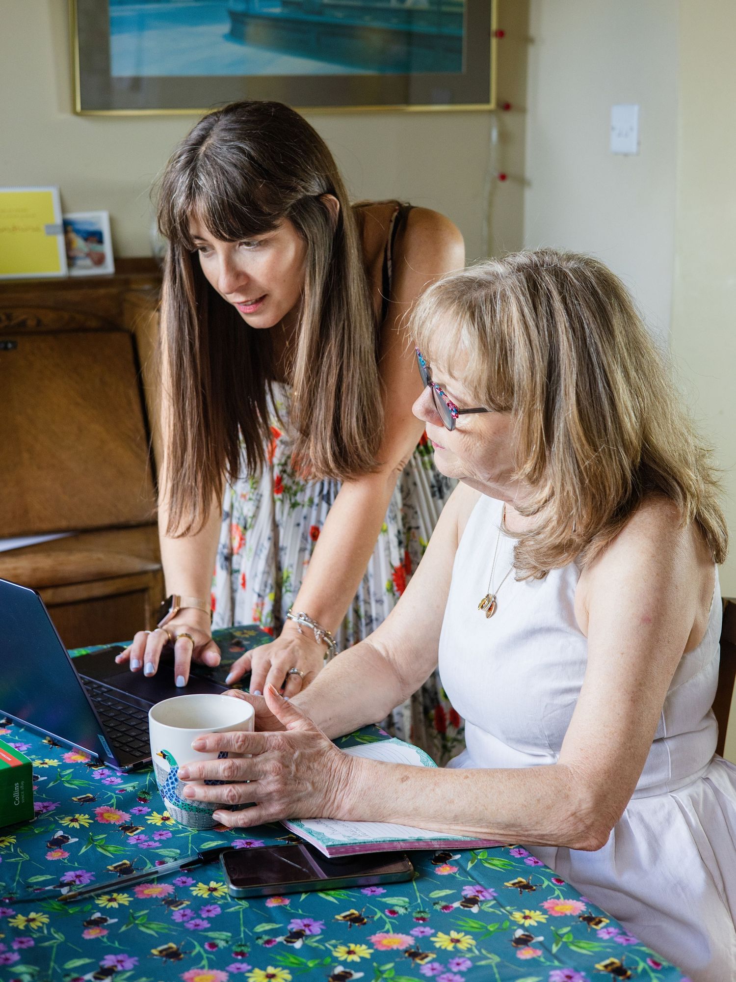 Two women collaborating on a laptop at a floral table, one standing and pointing at the screen while the other holds a mug.