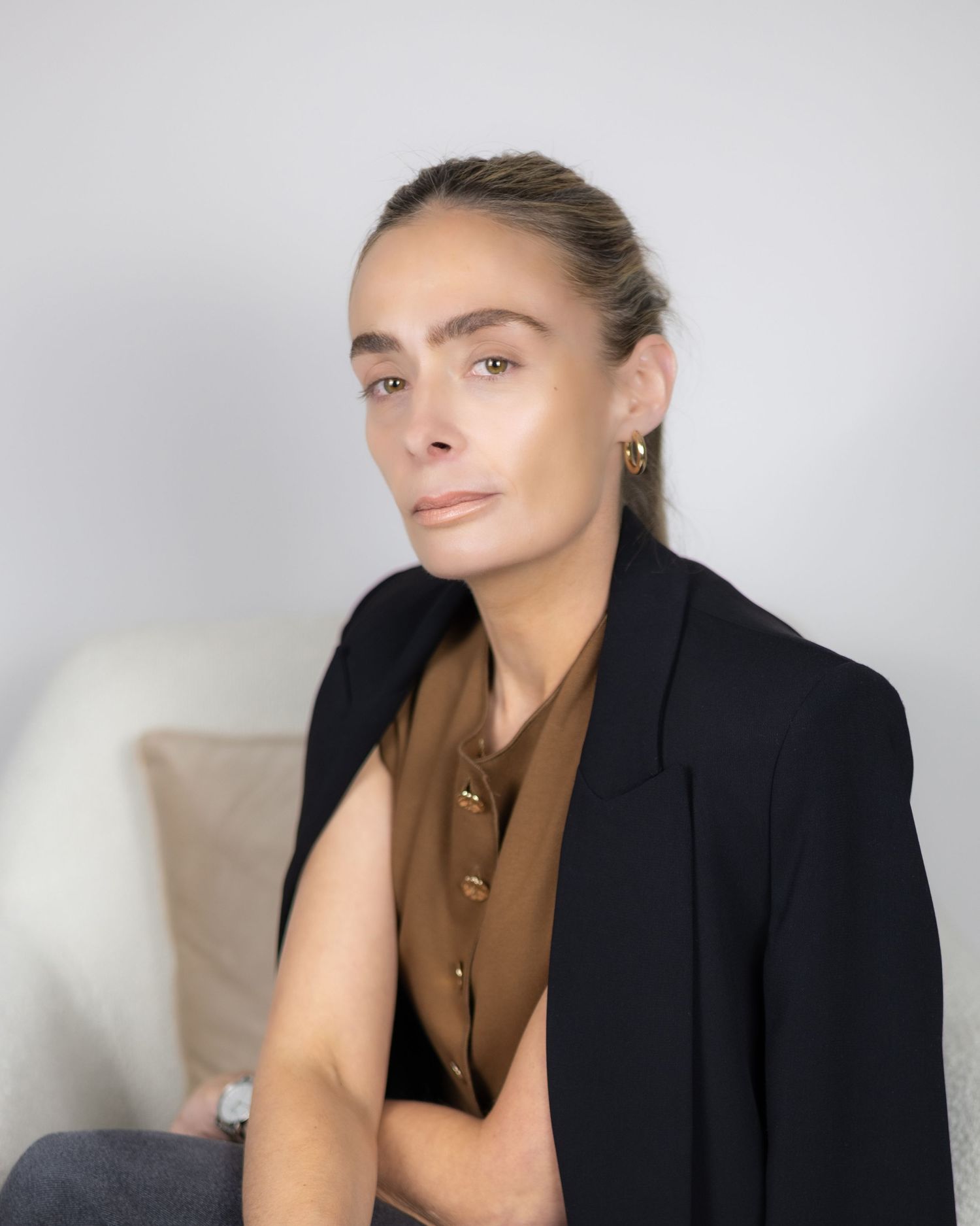 Elegant woman in black blazer and brown top with gold earrings sitting against a white background. Stylist in London