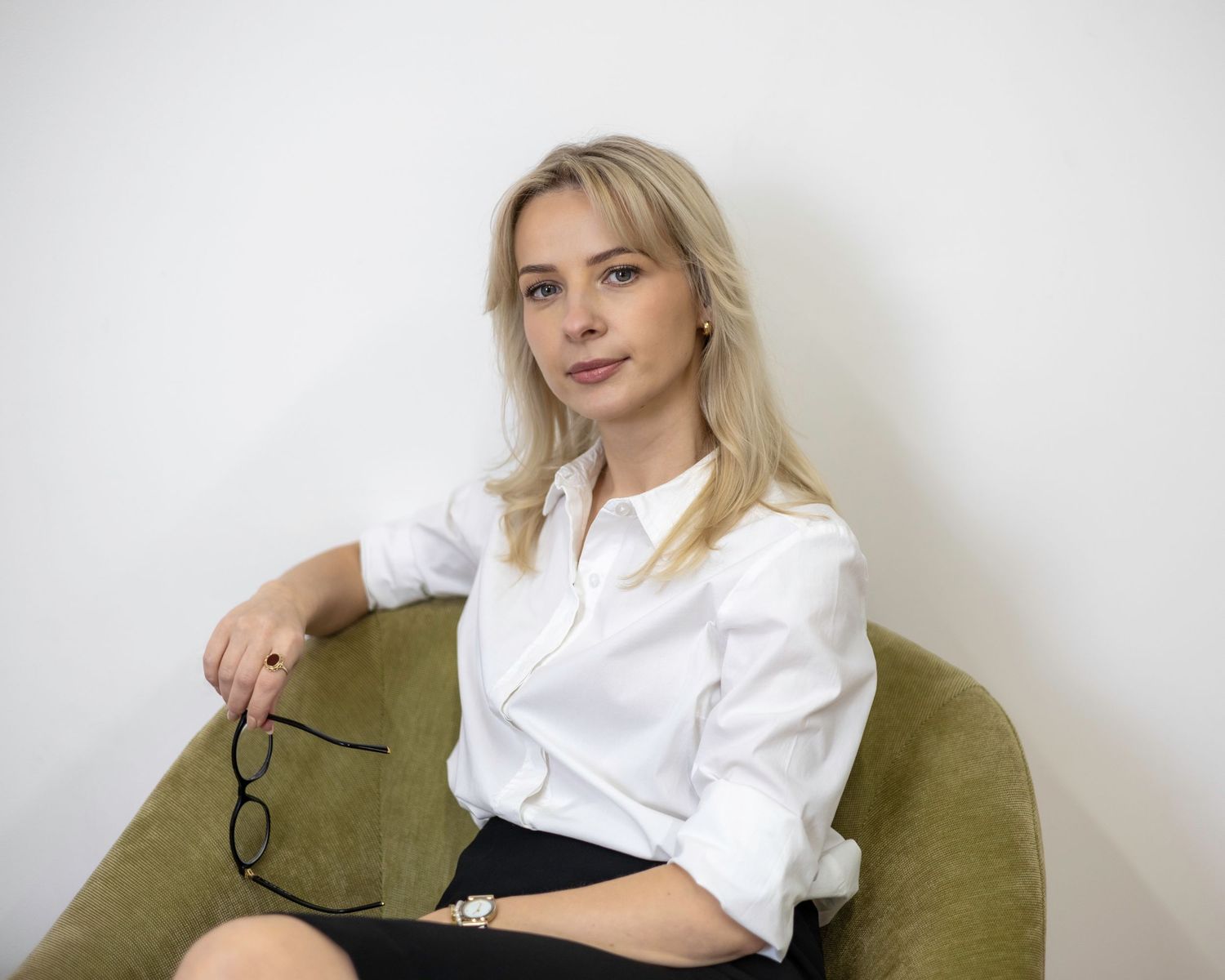 Professional blonde woman in white shirt sitting in olive green chair holding glasses. Brand Photography South London