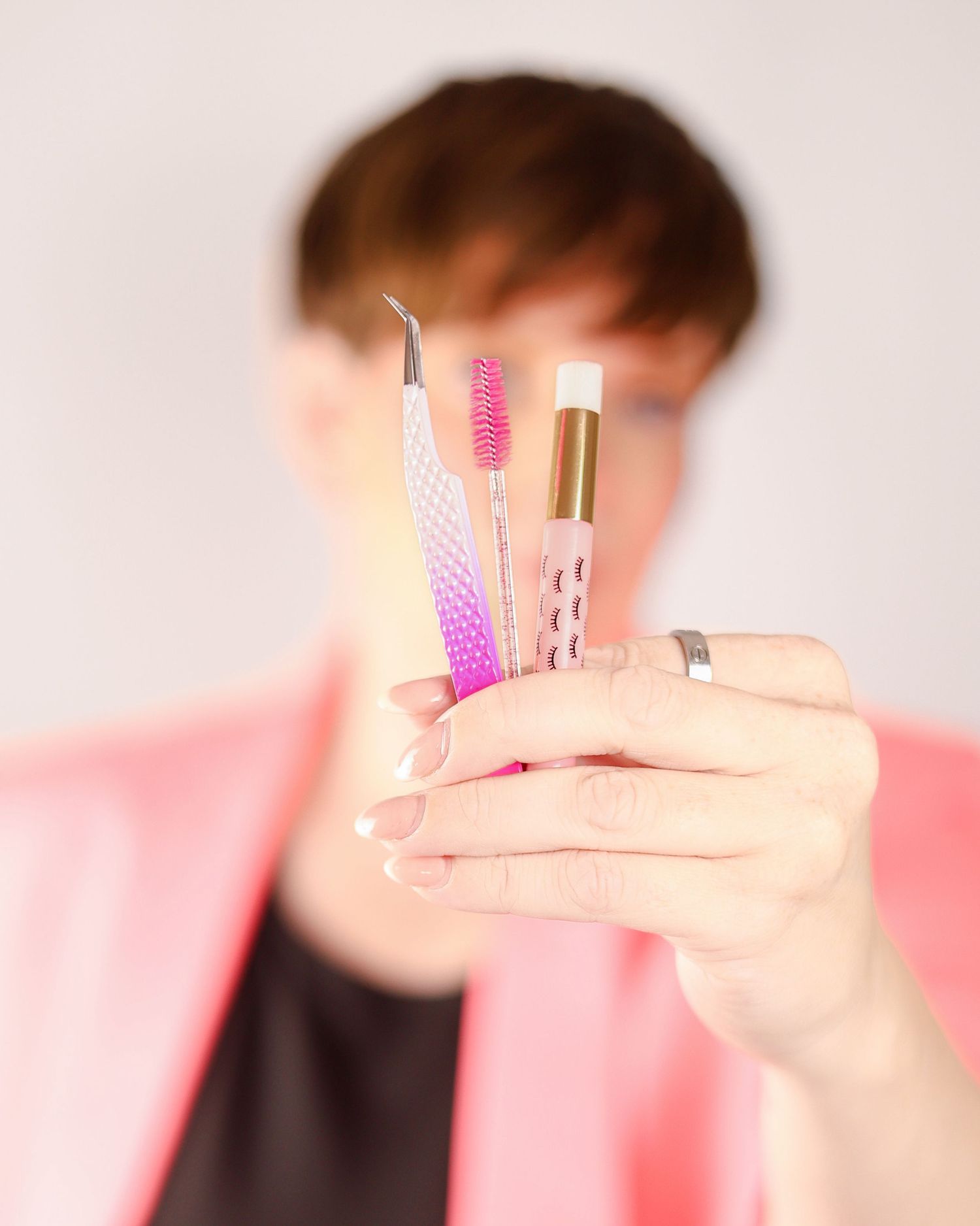 Woman in pink blazer holding pink tweezers and beauty tools toward camera, close-up. Selsdon, South London