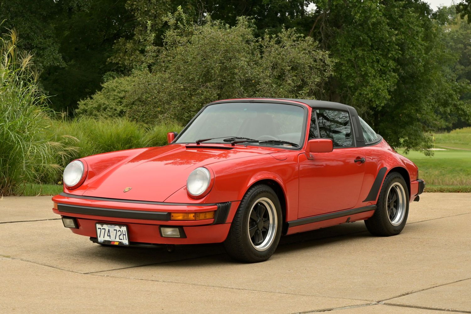 Red Porsche 911 Targa classic sports car with silver alloy wheels parked on a concrete driveway near green trees.