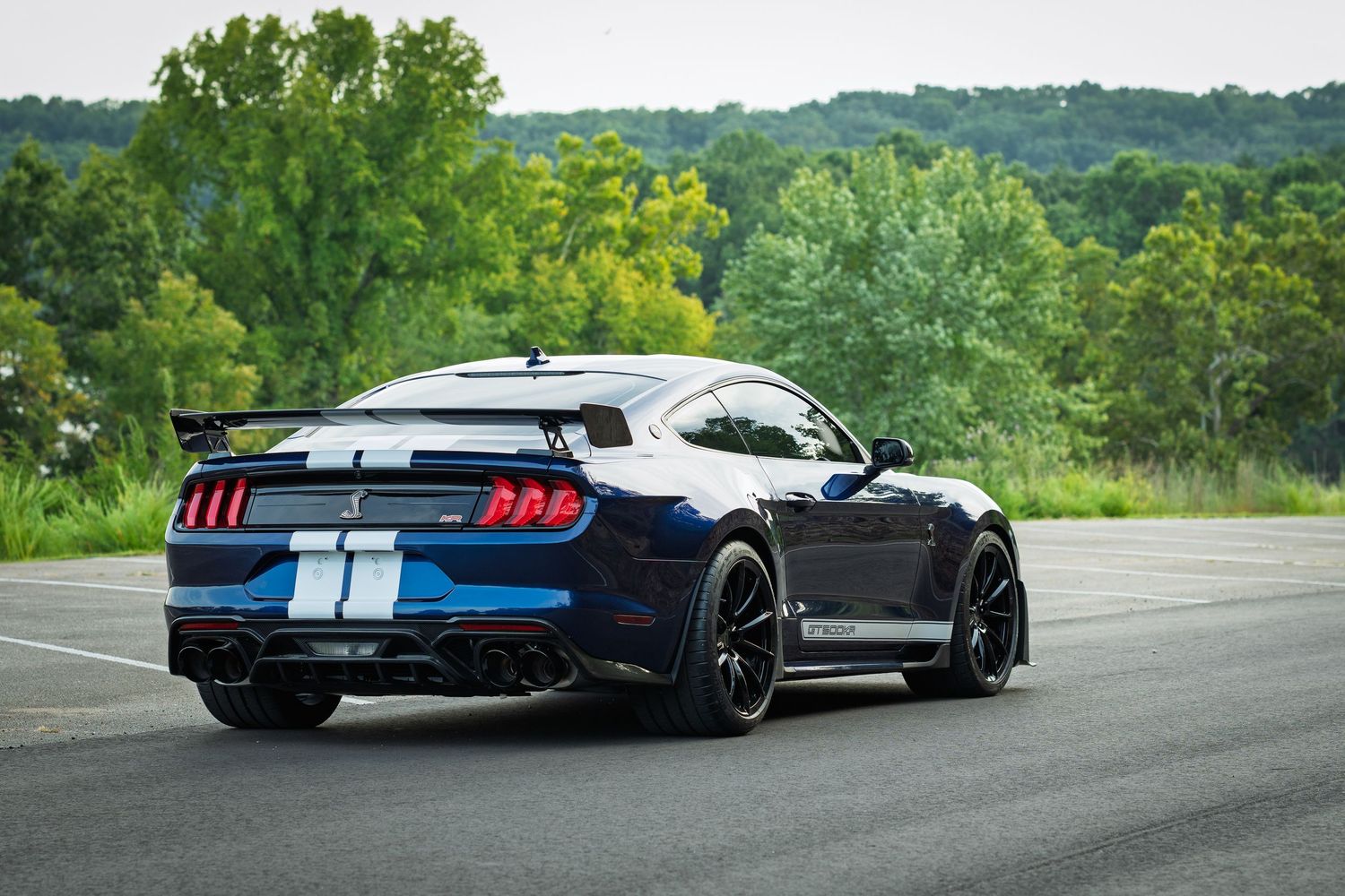 Blue Ford Shelby GT500 Mustang rear view on open road with white racing stripes and spoiler, surrounded by green trees.