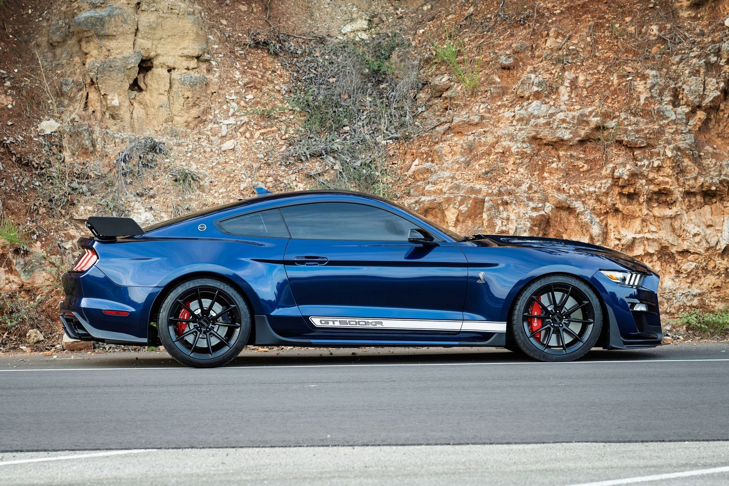 Blue Ford Shelby GT500 Mustang with white stripes parked beside rocky cliff side profile view.
