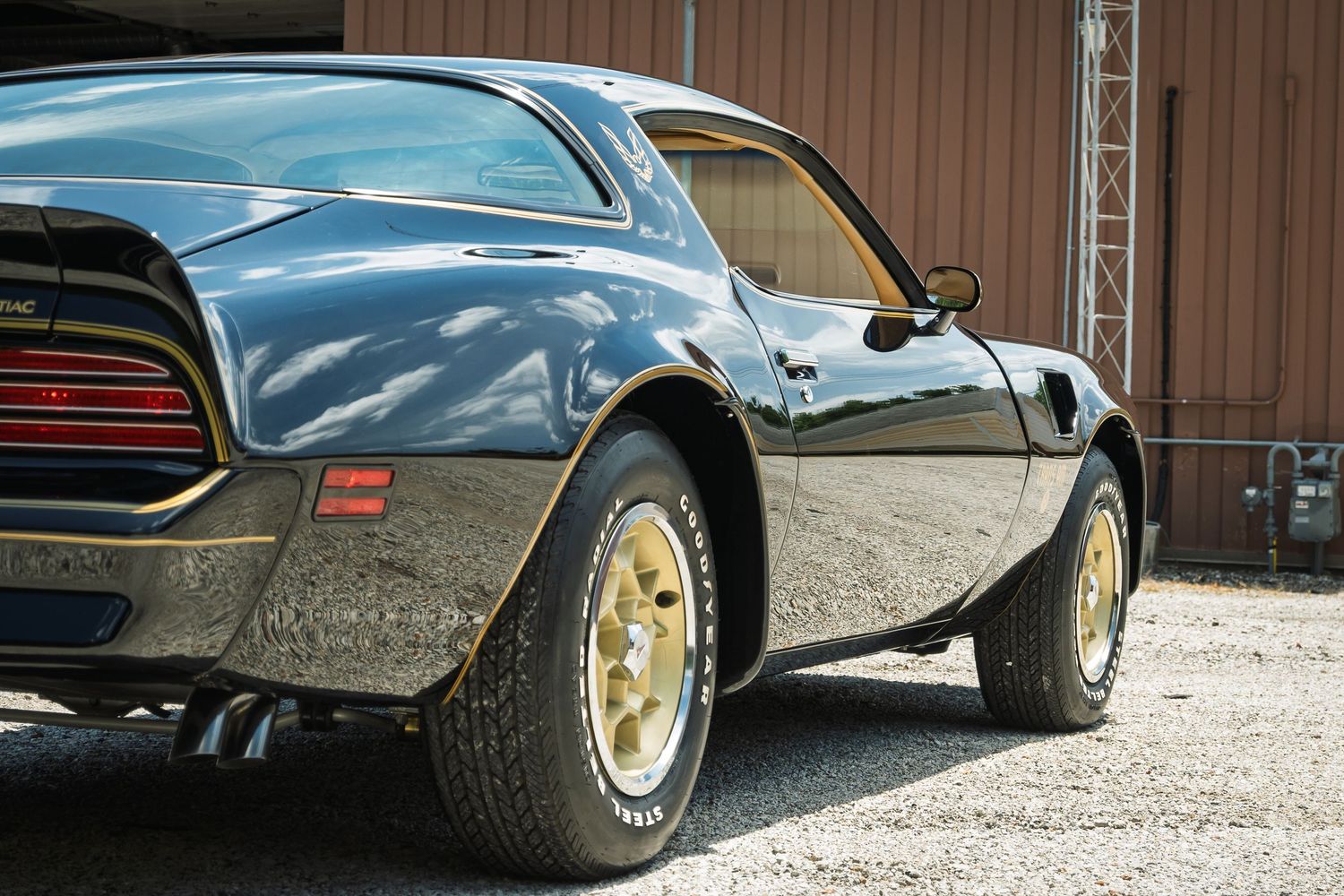 Rear view of classic black Pontiac Trans Am with gold wheels and dual exhaust on gravel lot.