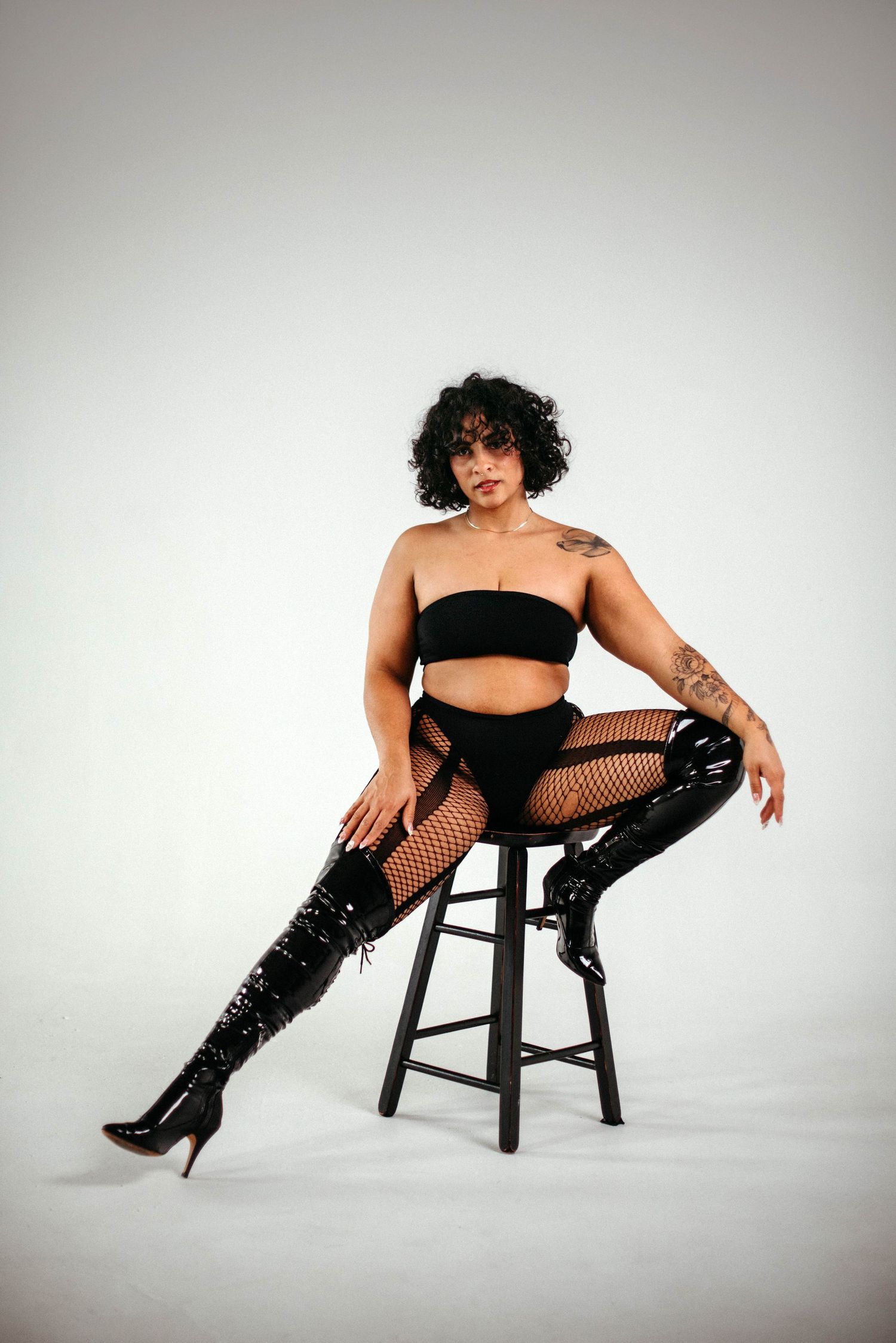 Woman in black lingerie, fishnet tights, and thigh-high boots posing on a stool in a studio setting.