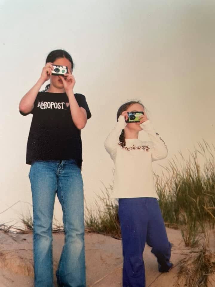 Two children photographing with cameras on a sandy beach with grass dunes in the background.