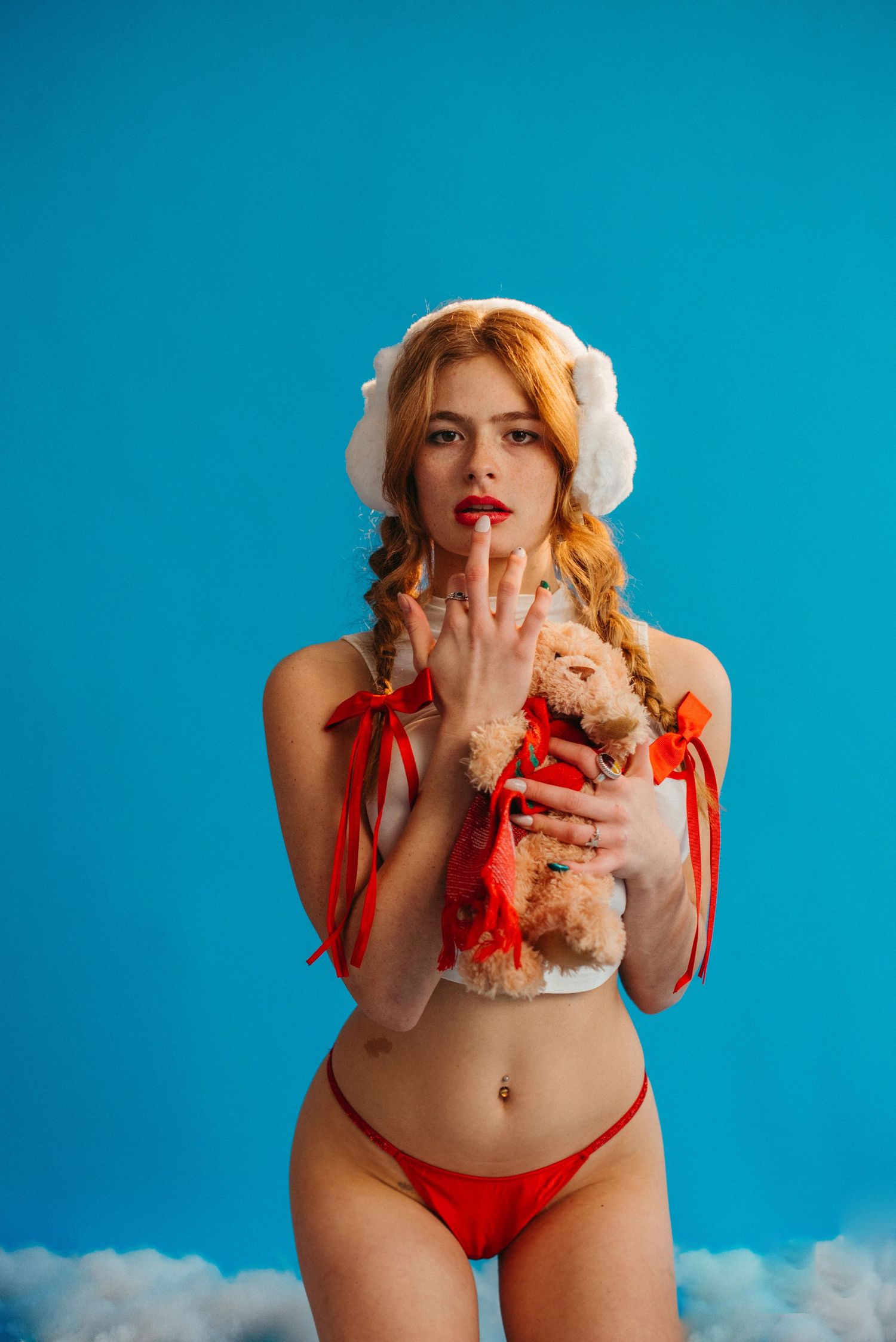 Young woman in red bikini and white earmuffs holding a teddy bear against a blue background.