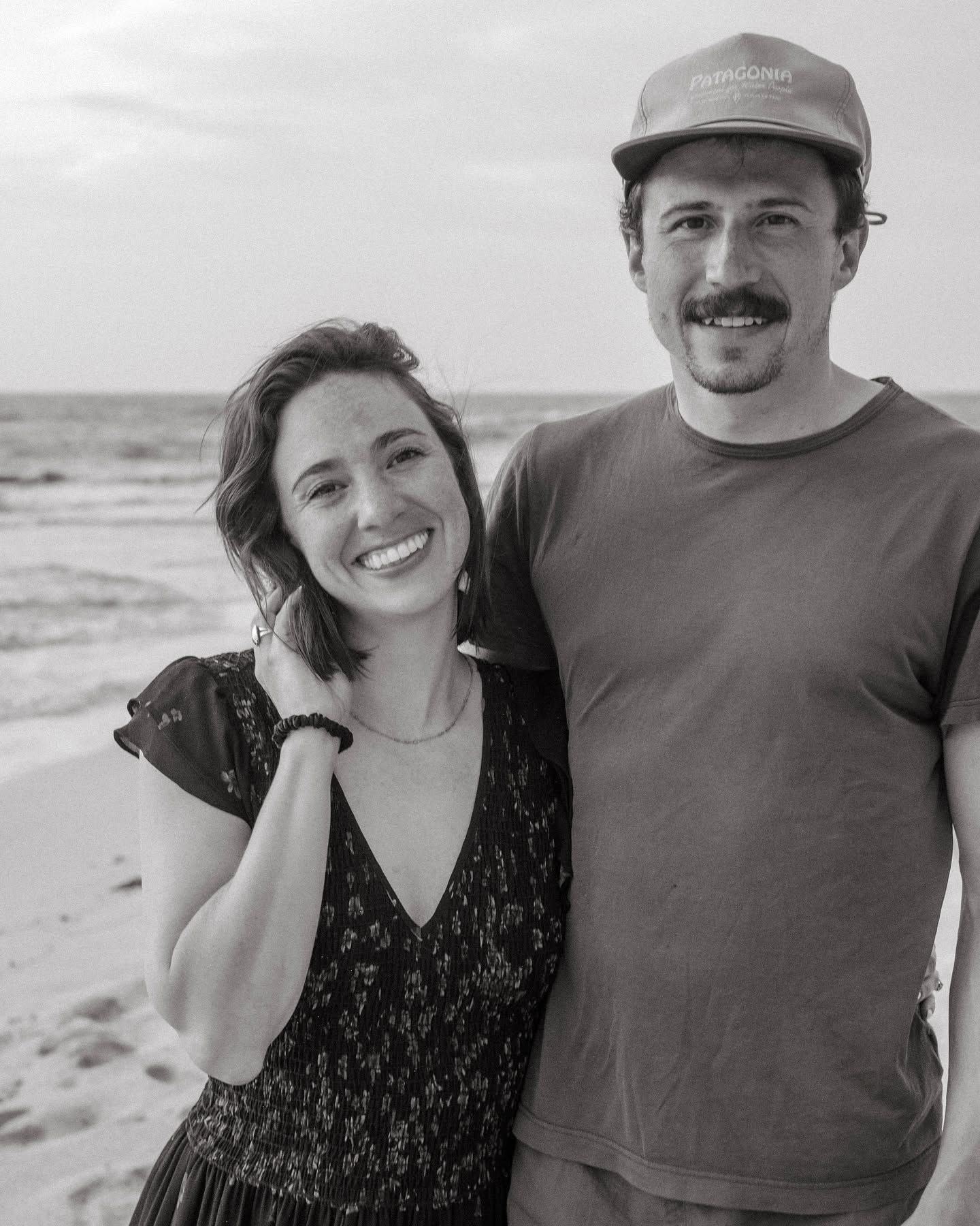 Black and white photo of a smiling couple posing together on a beach near the ocean.