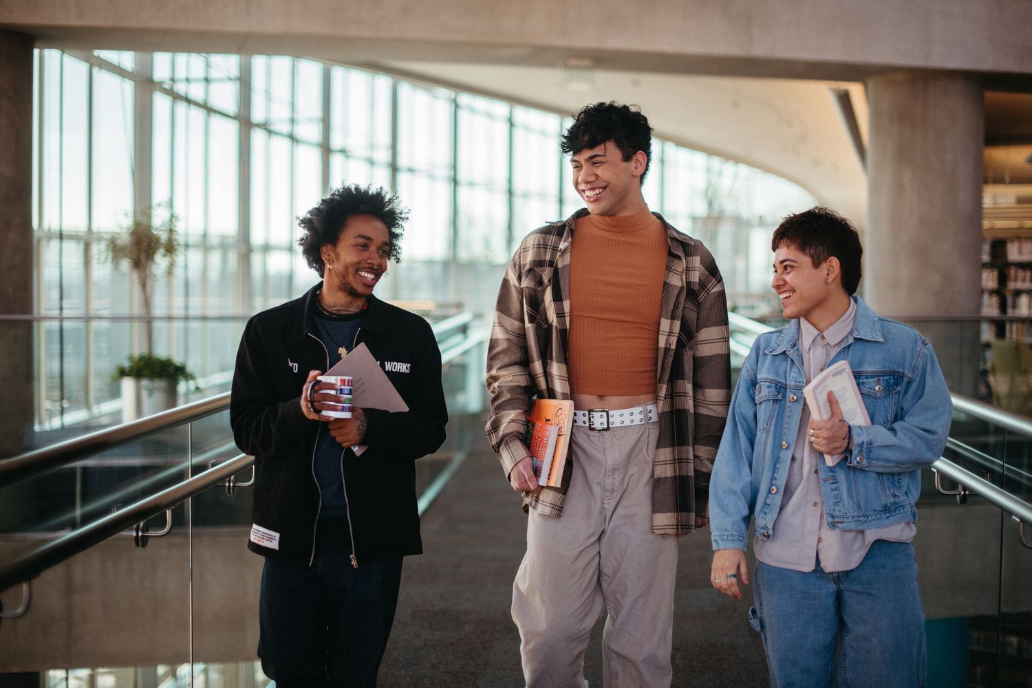 Three diverse college students walking and laughing together in a modern university building hallway.