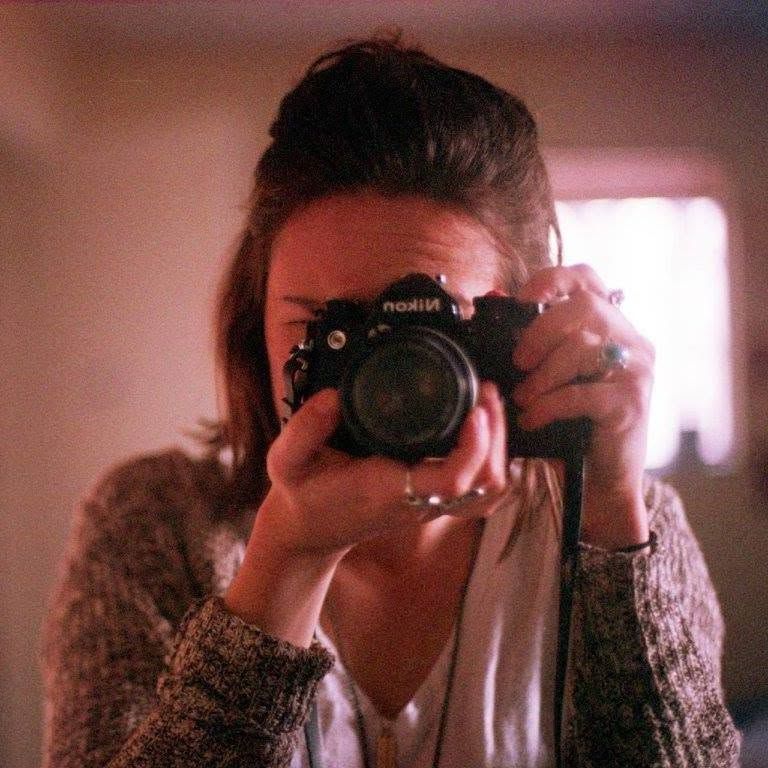 Young woman taking a self-portrait with a black Nikon film camera in a warmly lit room.