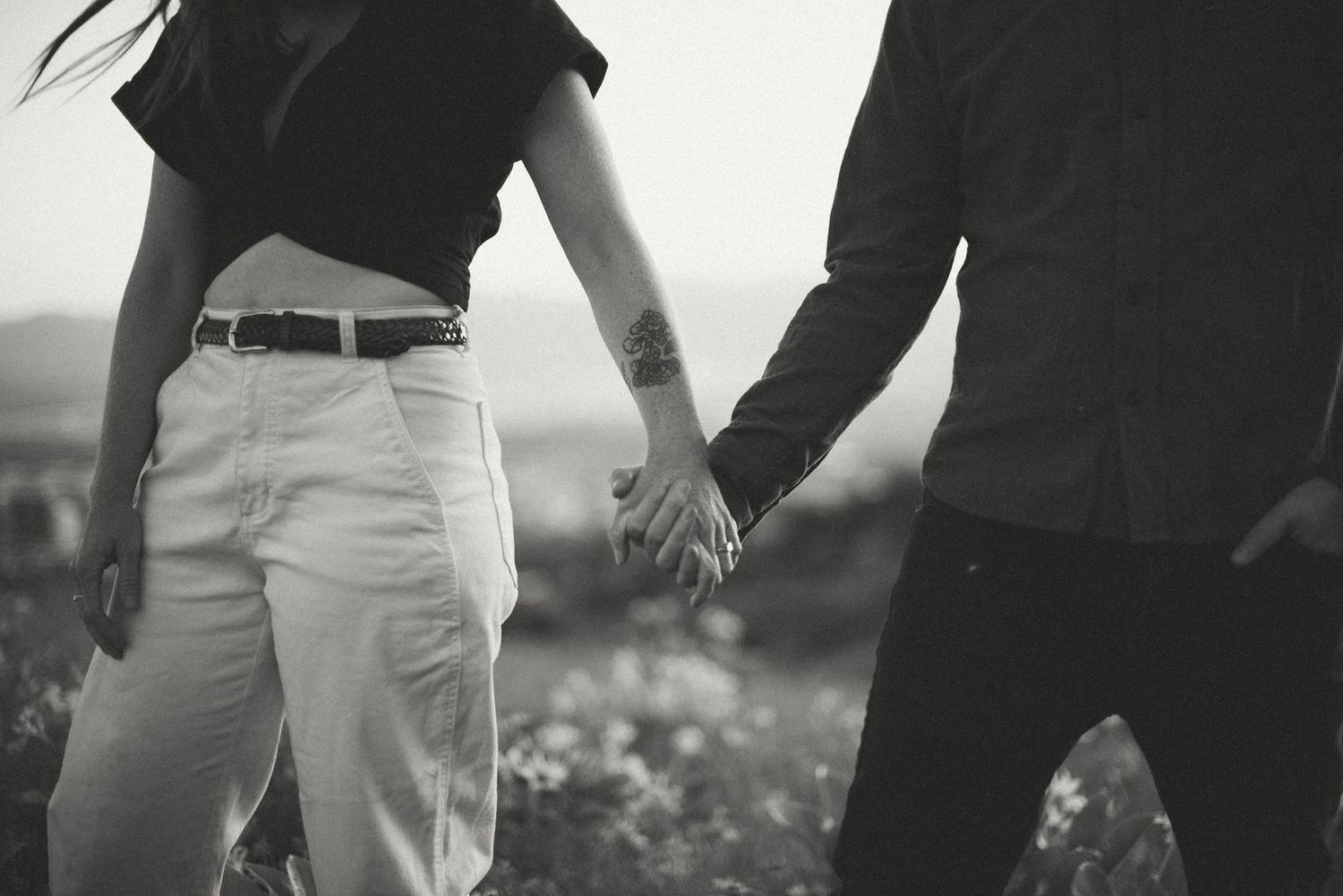 Black and white photo of a couple holding hands while walking through an open field.