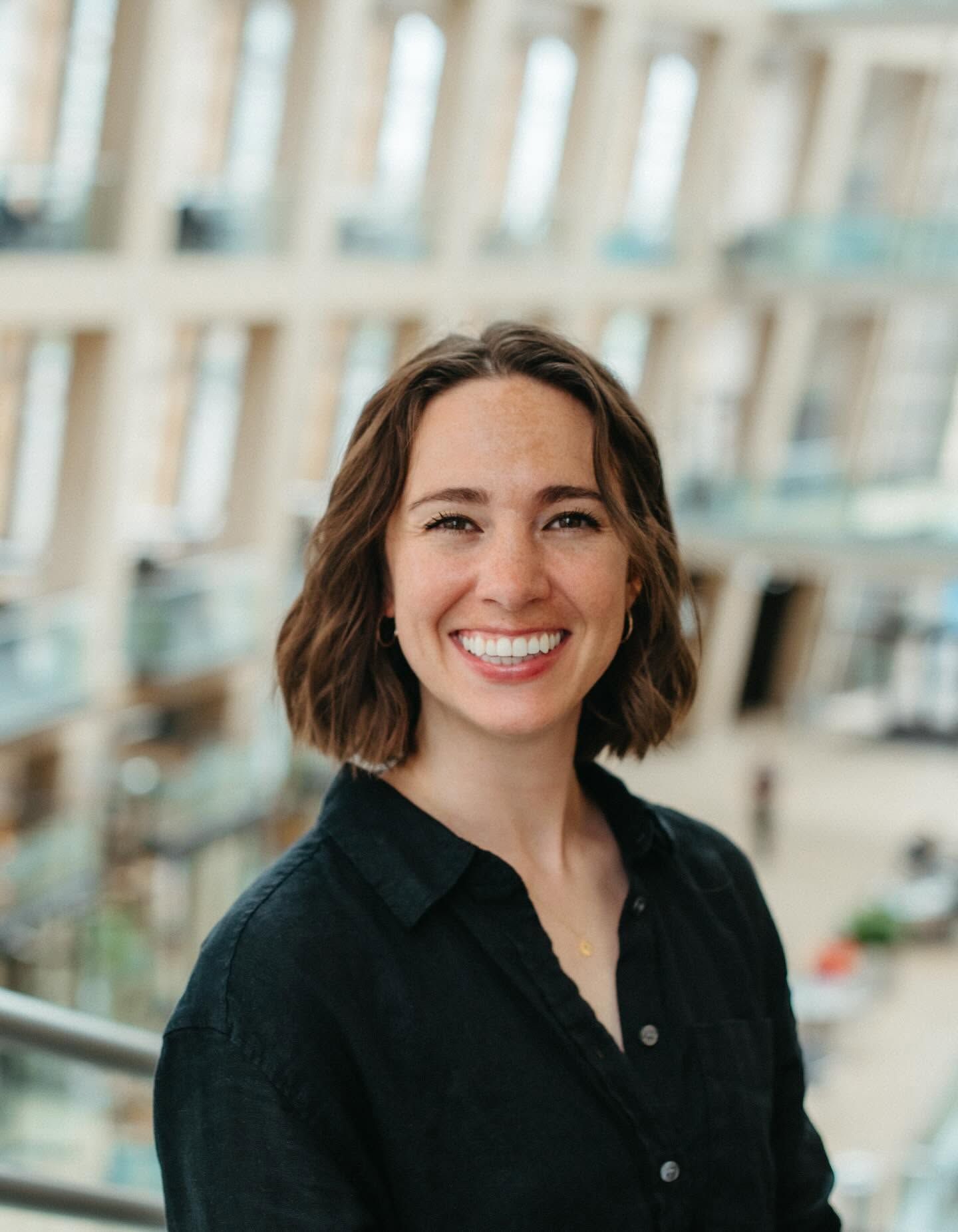 Smiling woman with short brown hair wearing a black shirt, posing in a modern building interior.