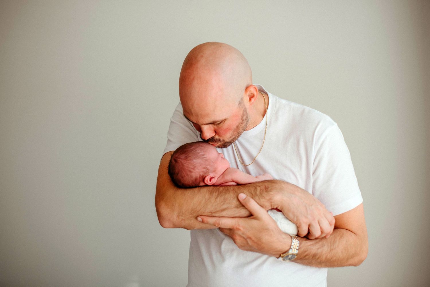 Bald father tenderly kissing his newborn baby while cradling the infant in his arms against a neutral background.