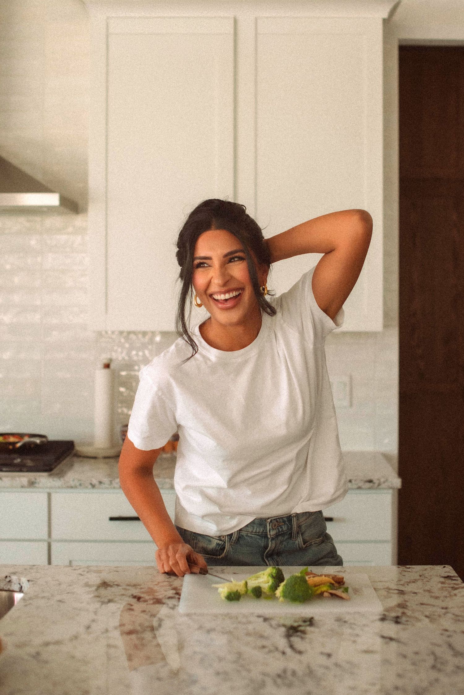 A laughing woman in a white t-shirt and jeans stands at a kitchen island with fresh vegetables.