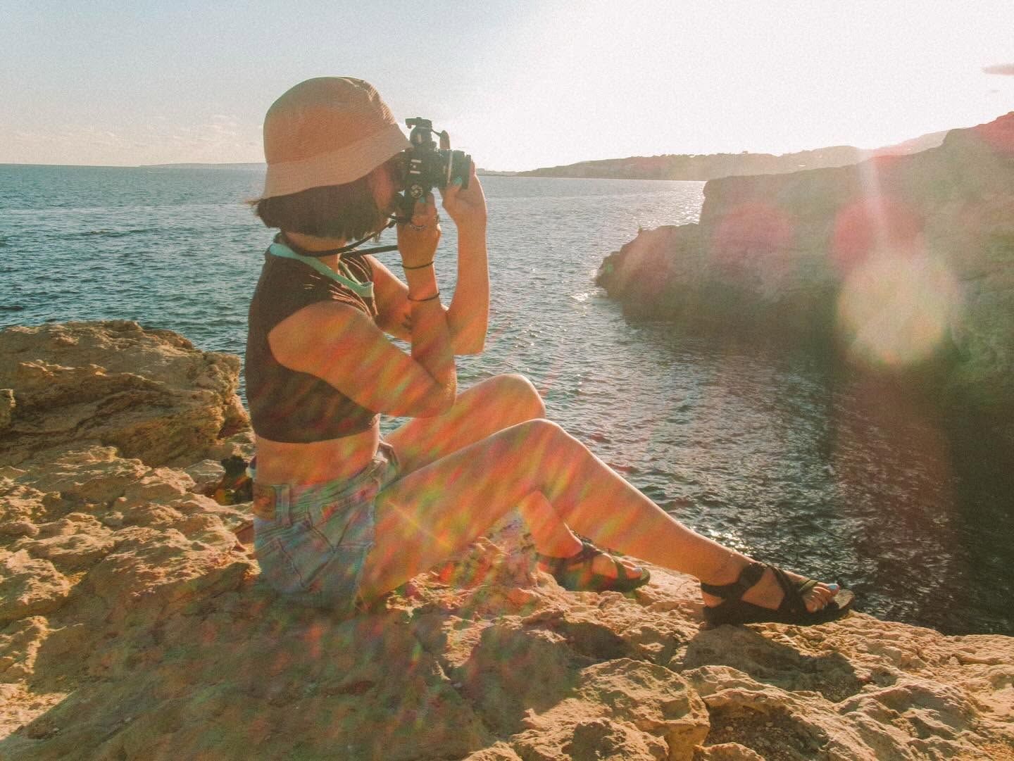 Woman sitting on rocky coastal cliffs photographing the sea at golden hour sunset with lens flare.