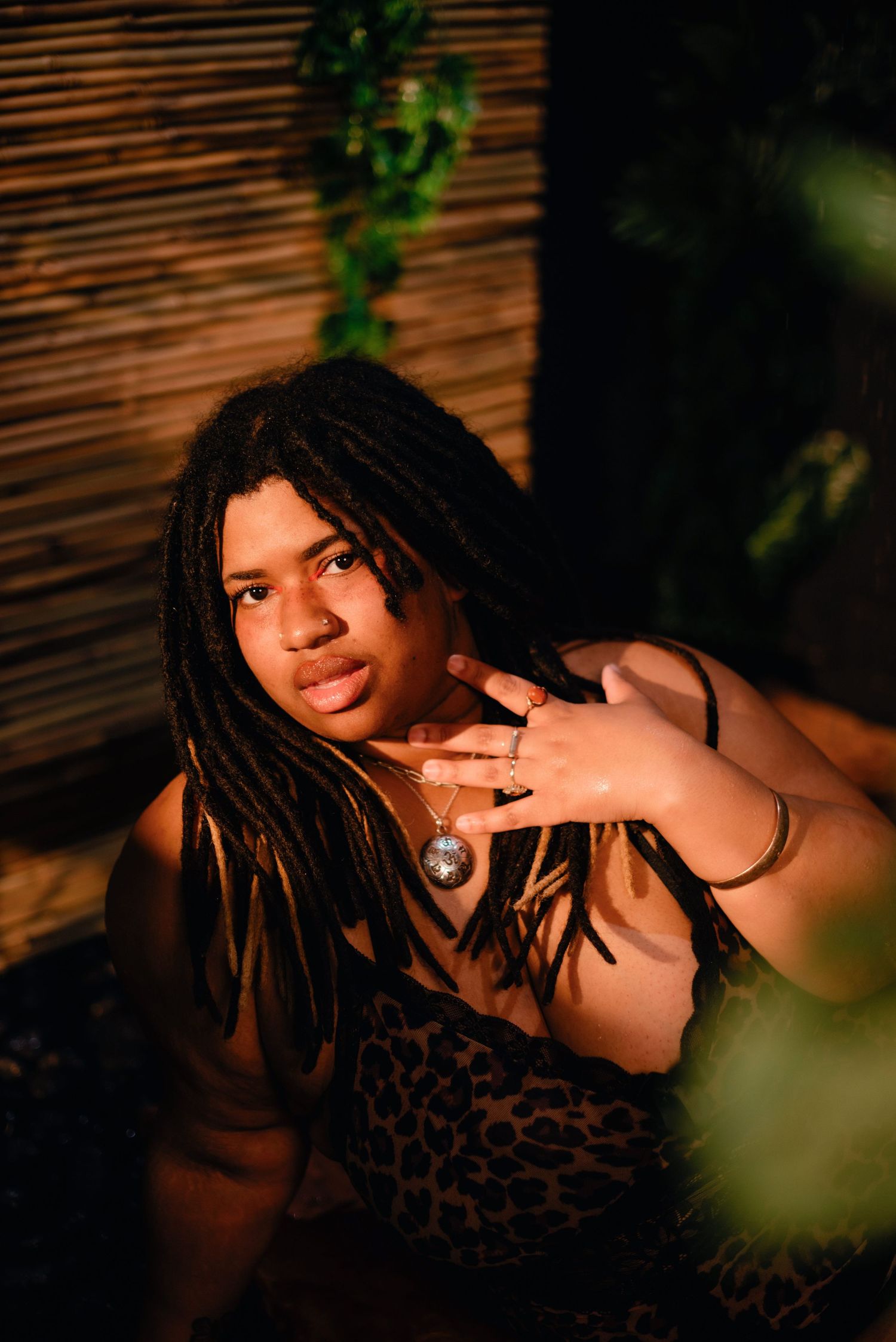 Young person with long dreadlocks wearing leopard print top and pendant necklace, posing outdoors in warm golden light.