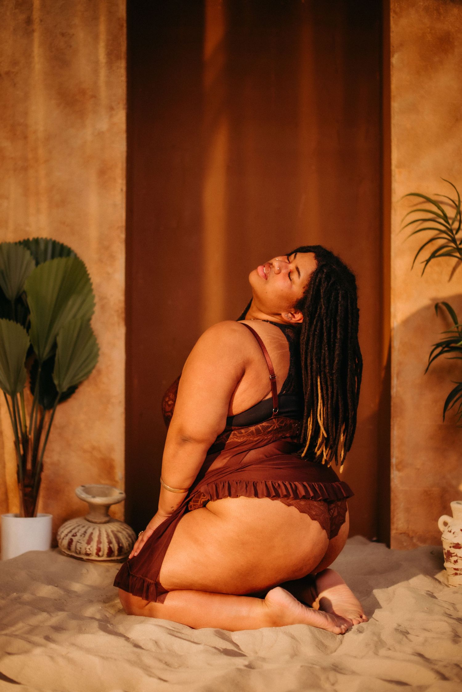Plus-size woman with locs poses gracefully on sandy floor in brown lingerie, surrounded by tropical plants in warm light.