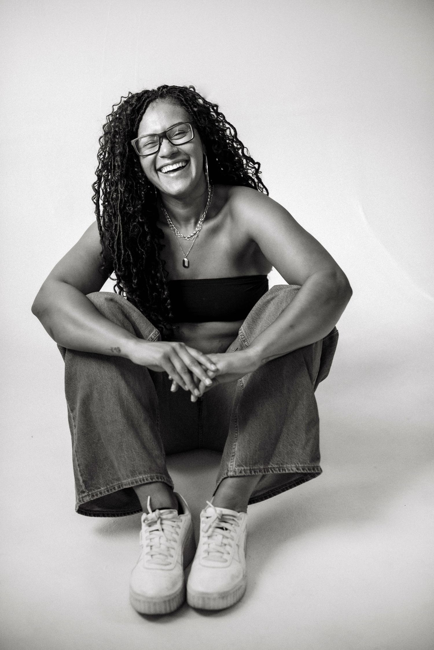 Black and white photo of a laughing woman with curly hair, glasses, sitting cross-legged in jeans and sneakers.