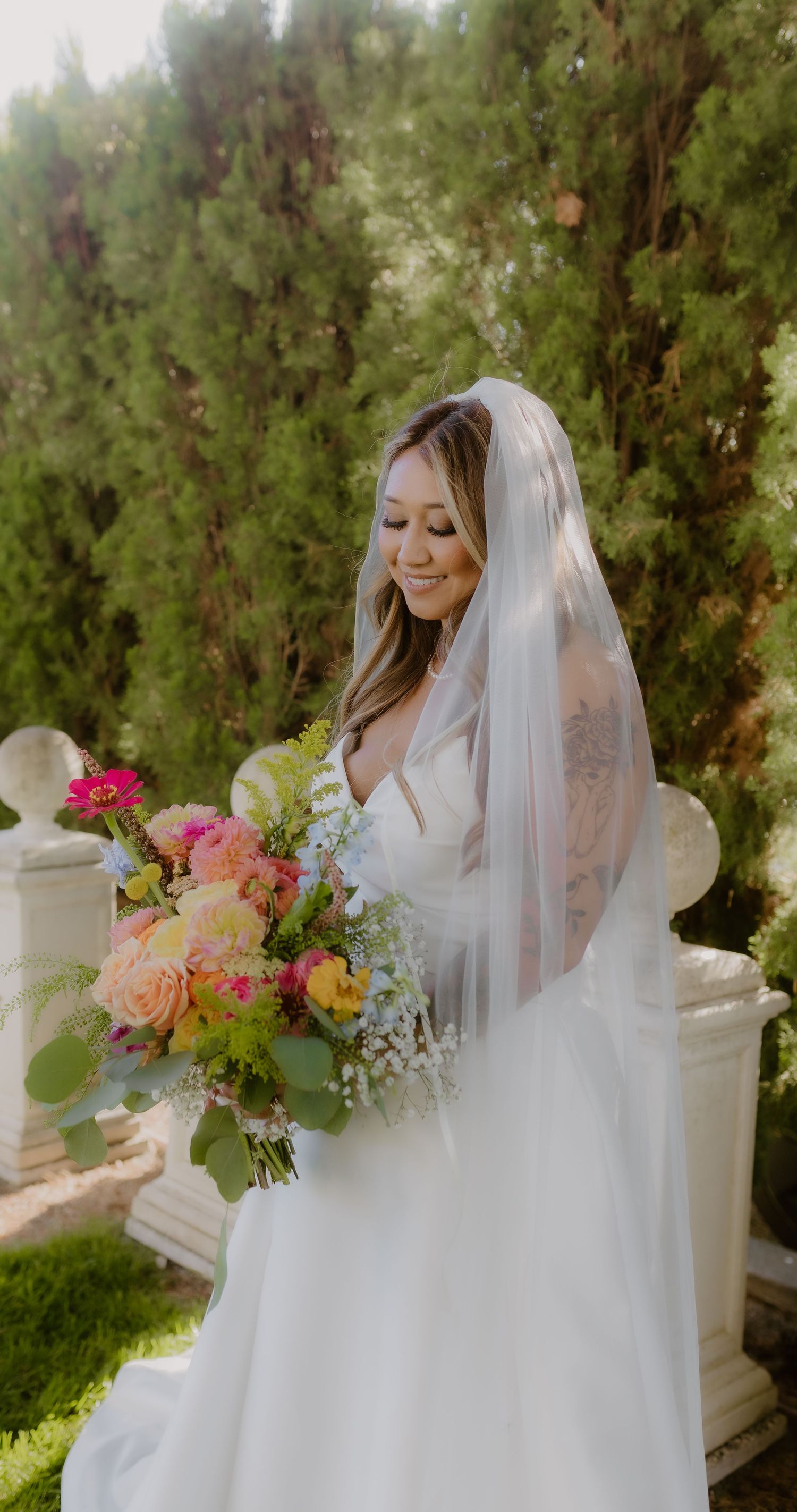 Radiant bride in white gown and veil smiles at colorful bridal bouquet in a garden setting.