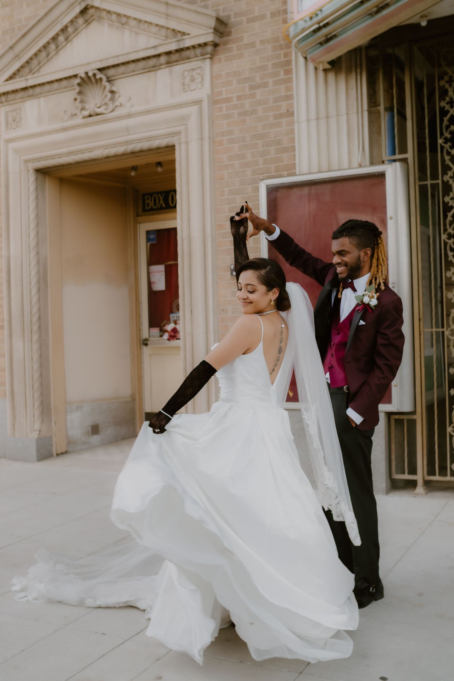Joyful groom in burgundy tuxedo twirling bride in flowing white gown outside historic Warner's Theatre wedding venue.