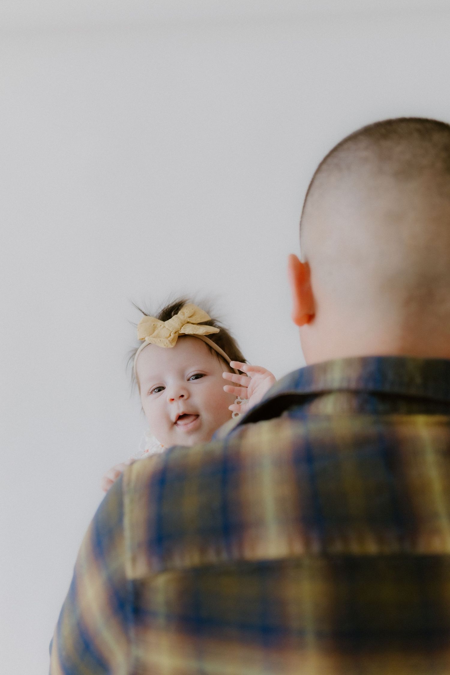 A father in plaid shirt holds his newborn baby girl with yellow bow headband against white background.