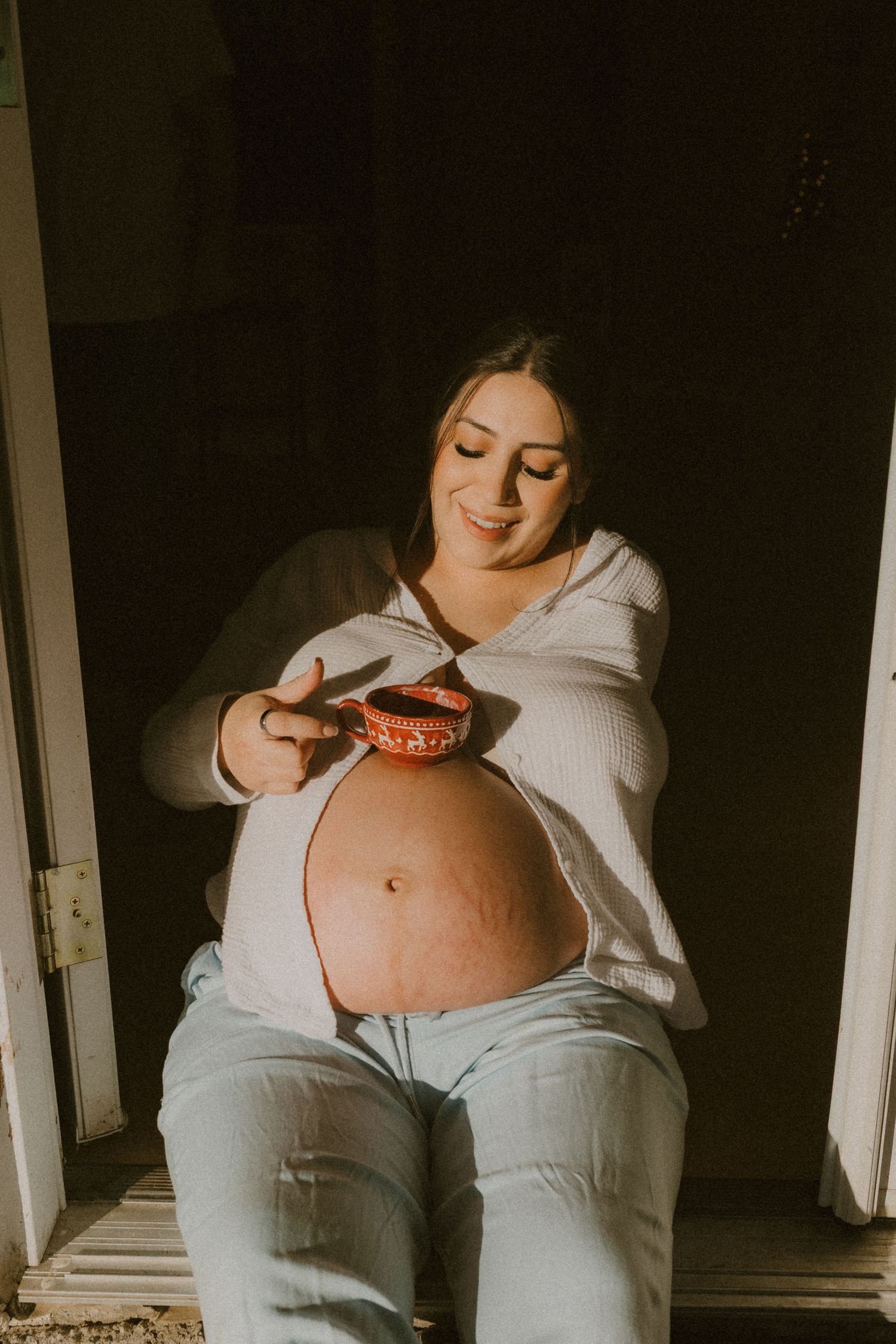 Pregnant woman in white top sits in golden sunlight, smiling while holding a decorative red mug near belly.