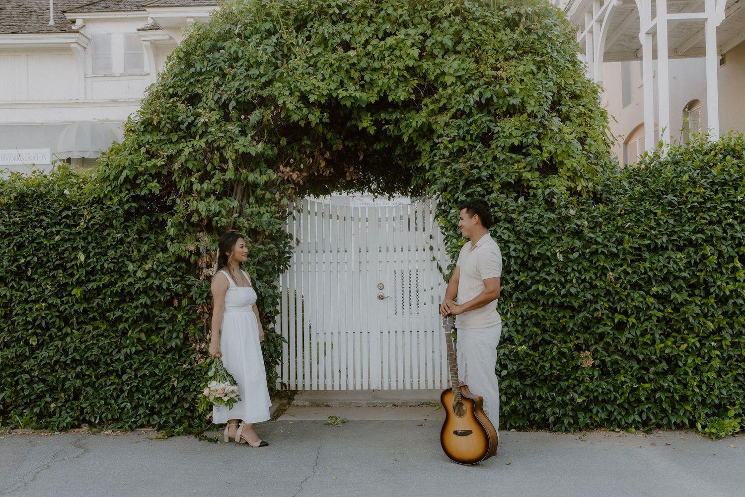 Couple in white outfits facing each other by ivy-covered gate arch with acoustic guitar.