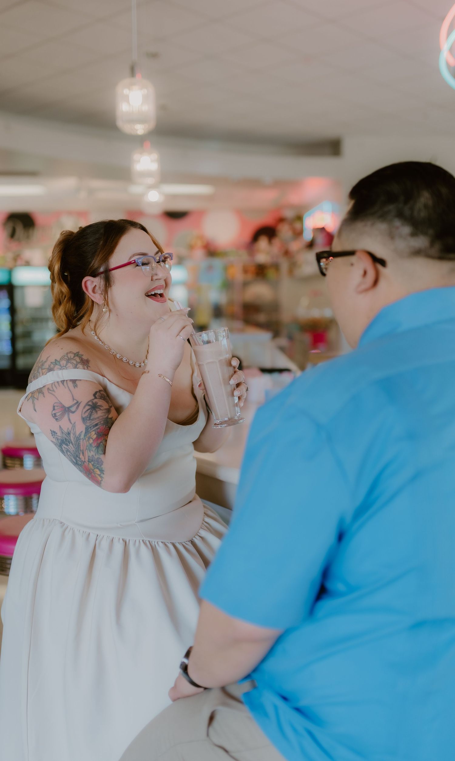 Tattooed bride in white dress laughing while sipping milkshake with groom in blue shirt at retro diner.