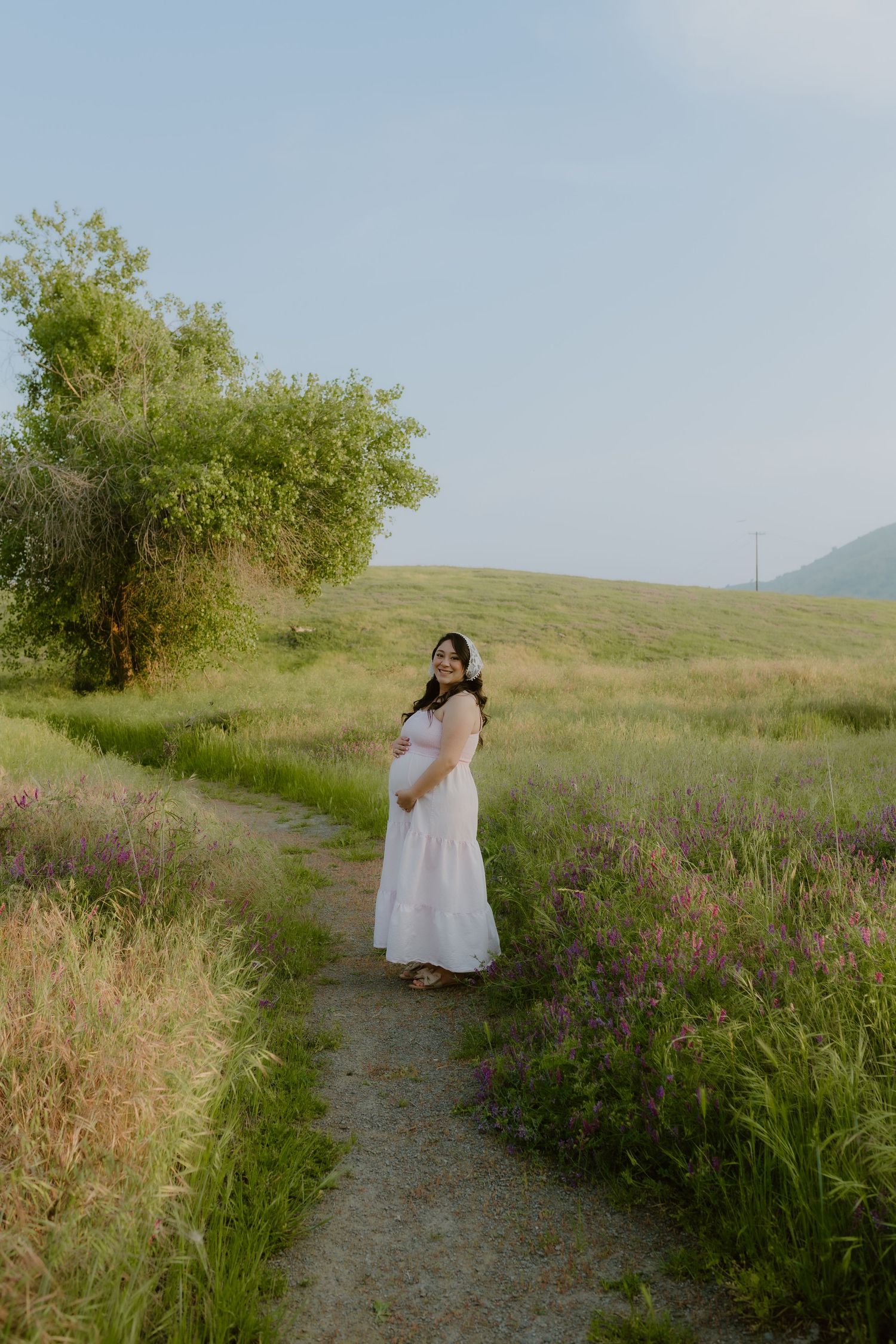 A pregnant woman in a white dress cradles her belly while standing on a scenic wildflower meadow path.