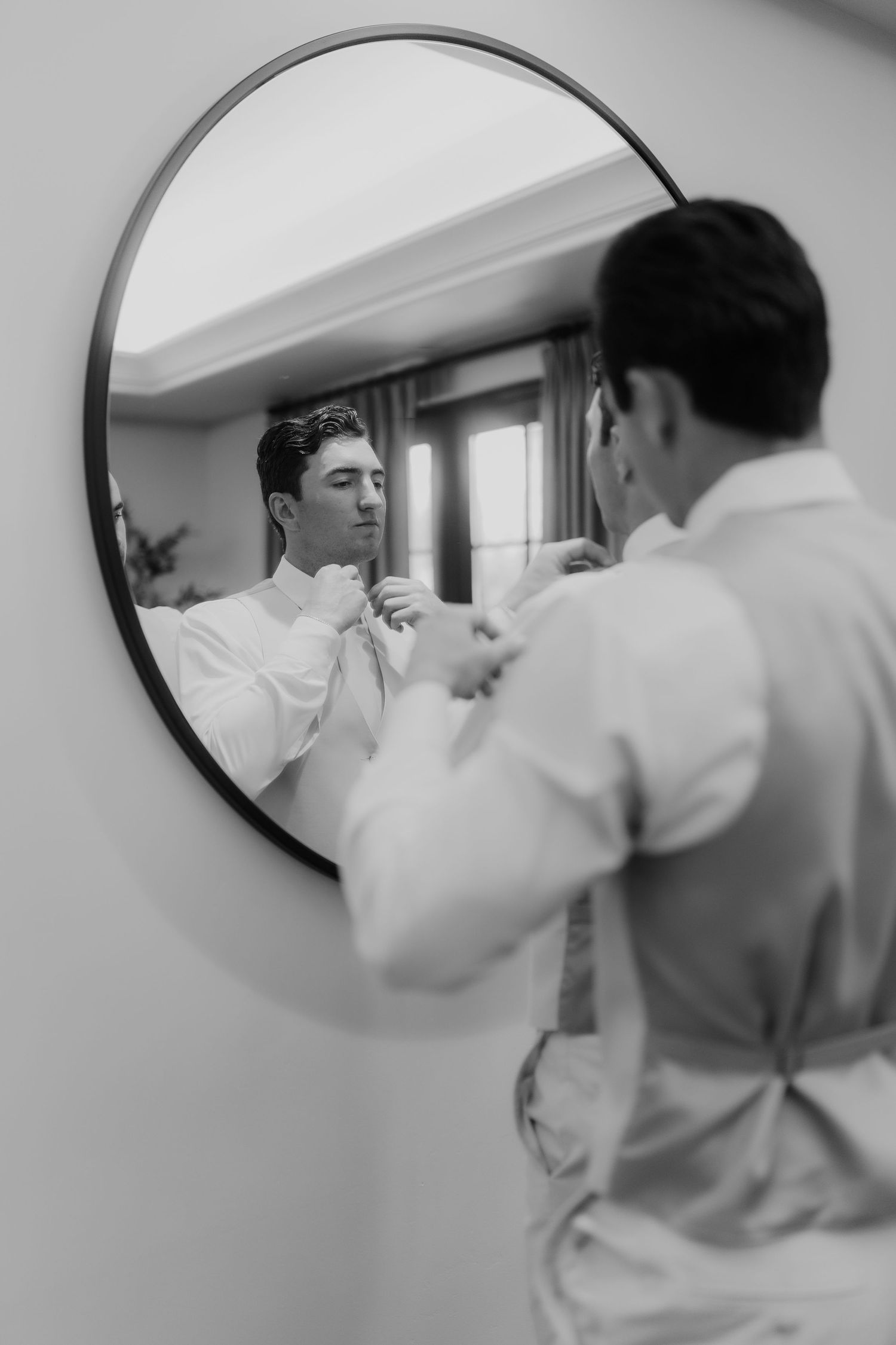 Black and white photo of groom adjusting his tie while looking into a round mirror.