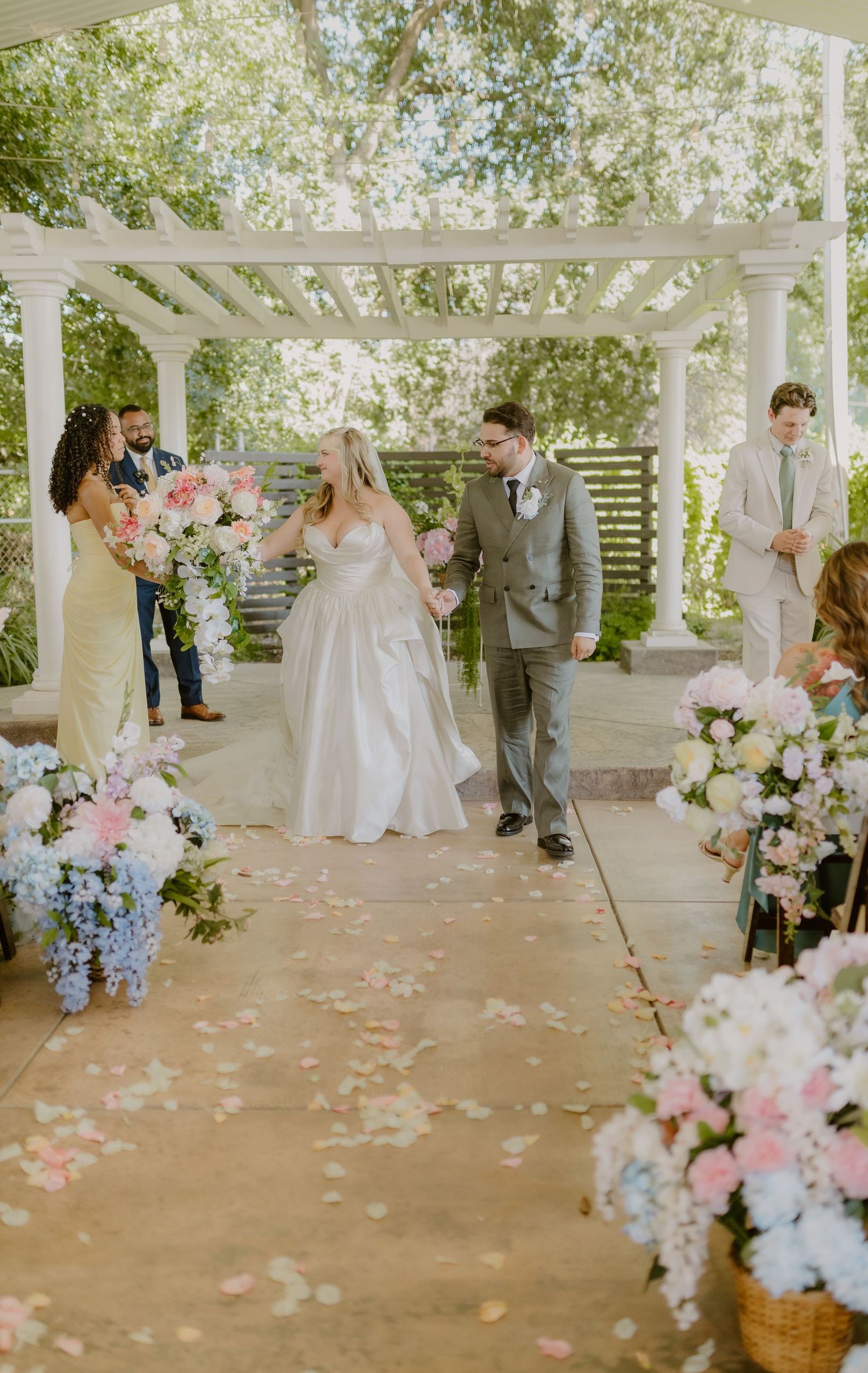 Newlyweds walking down petal-lined aisle surrounded by pink and blue floral arrangements.