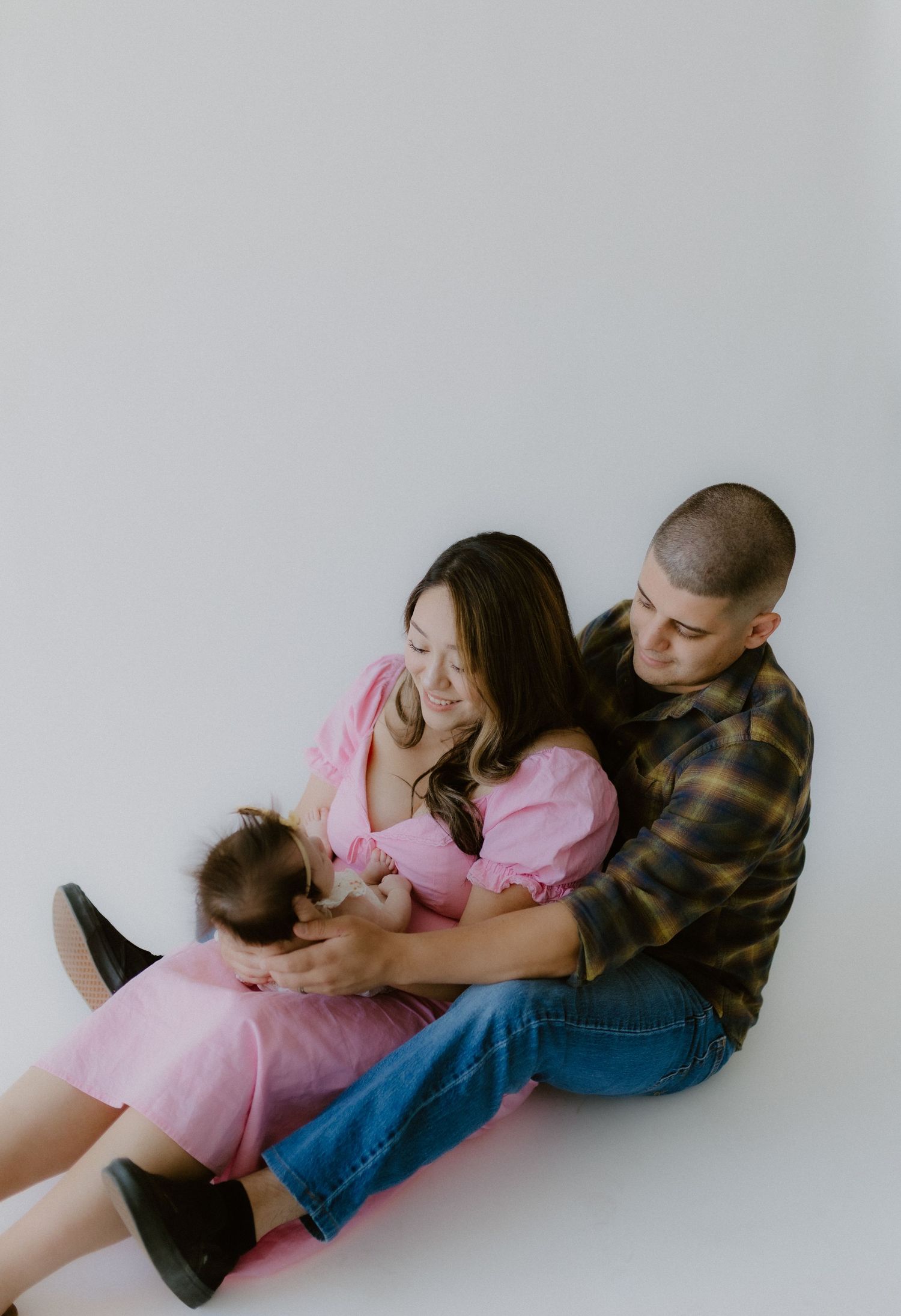 A couple sits together on a white floor, the mother in pink cradling their newborn baby.
