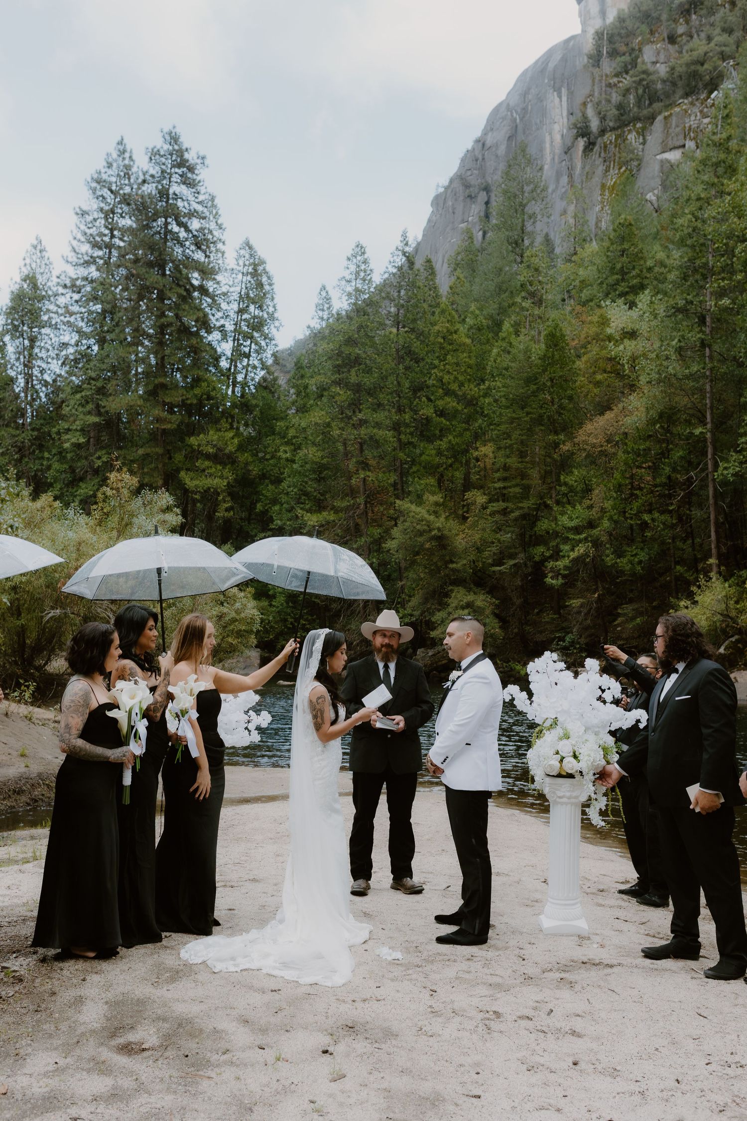 Outdoor wedding ceremony in Yosemite with bride, groom, and wedding party holding clear umbrellas in the rain.