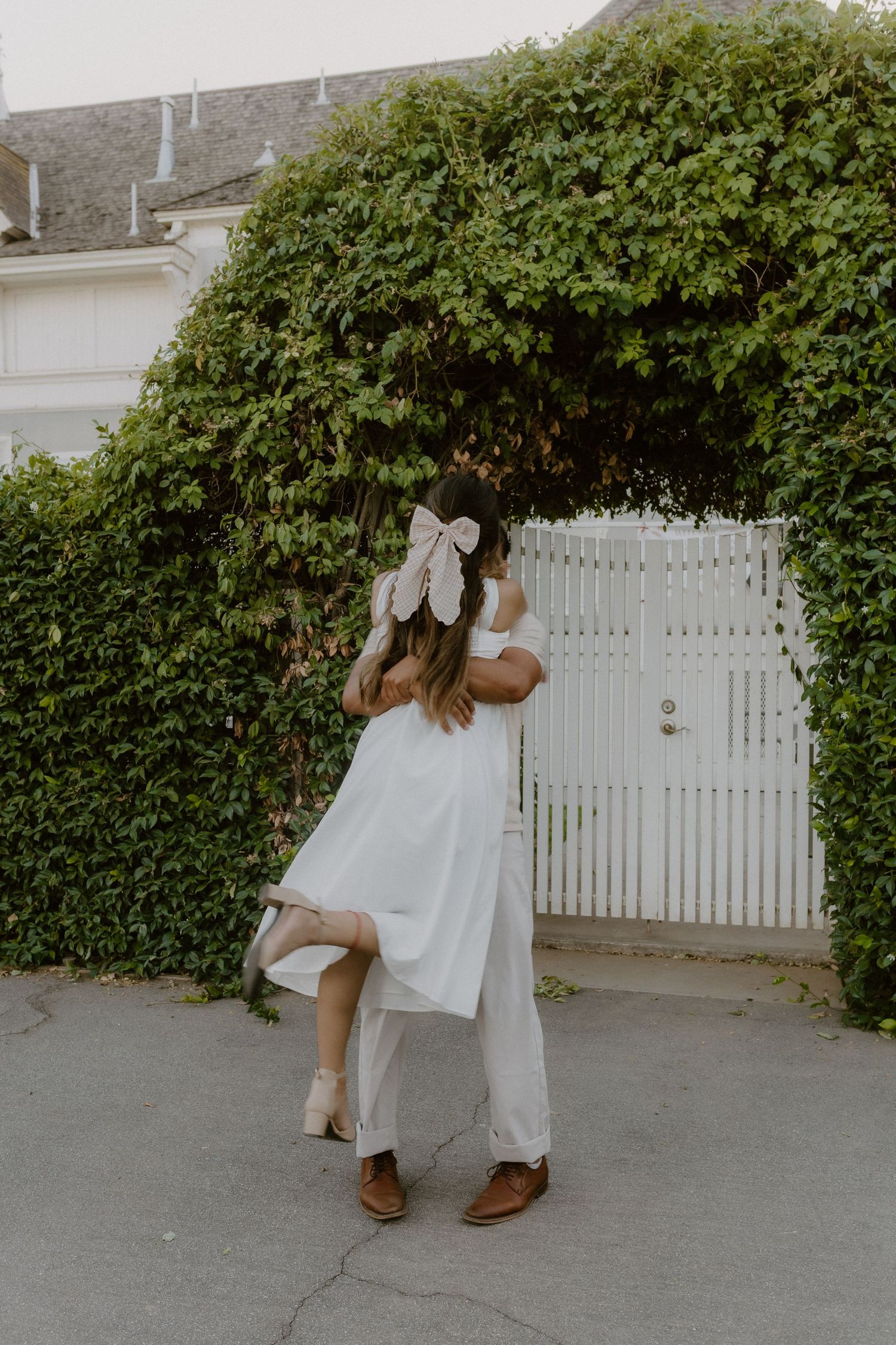 Woman in white dress with large bow being lifted by partner near ivy-covered white gate.