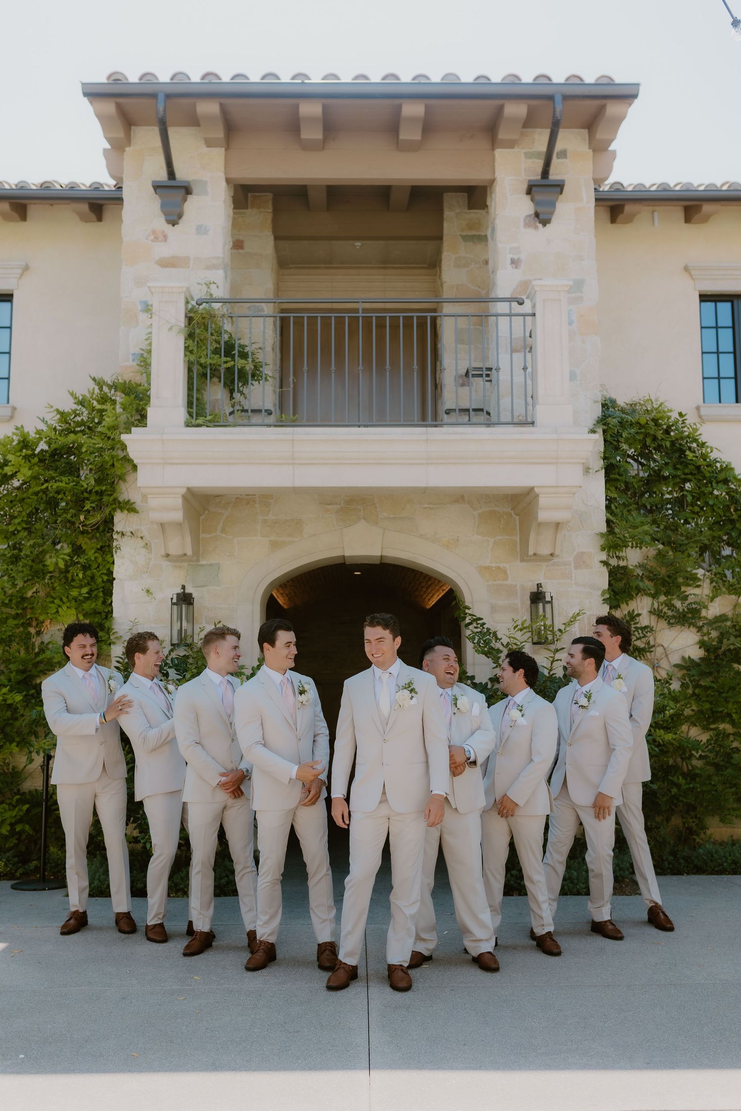 Groom and groomsmen in matching beige suits posing in front of elegant stone villa wedding venue. Monserate Winery, Fallbrook, California