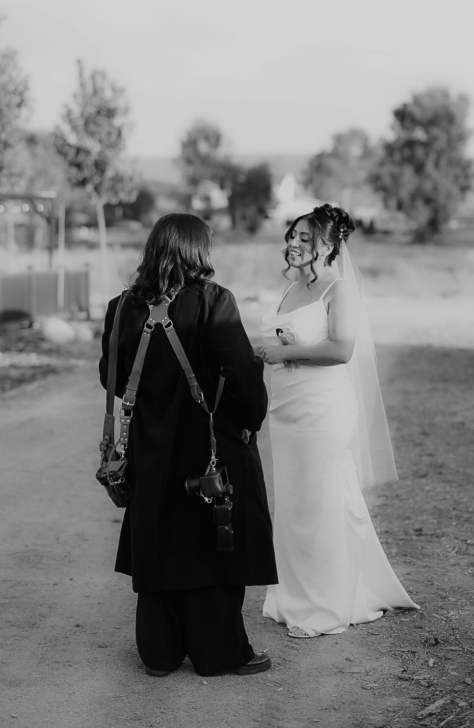Black and white photo of wedding photographer in black coat speaking with smiling bride outdoors.