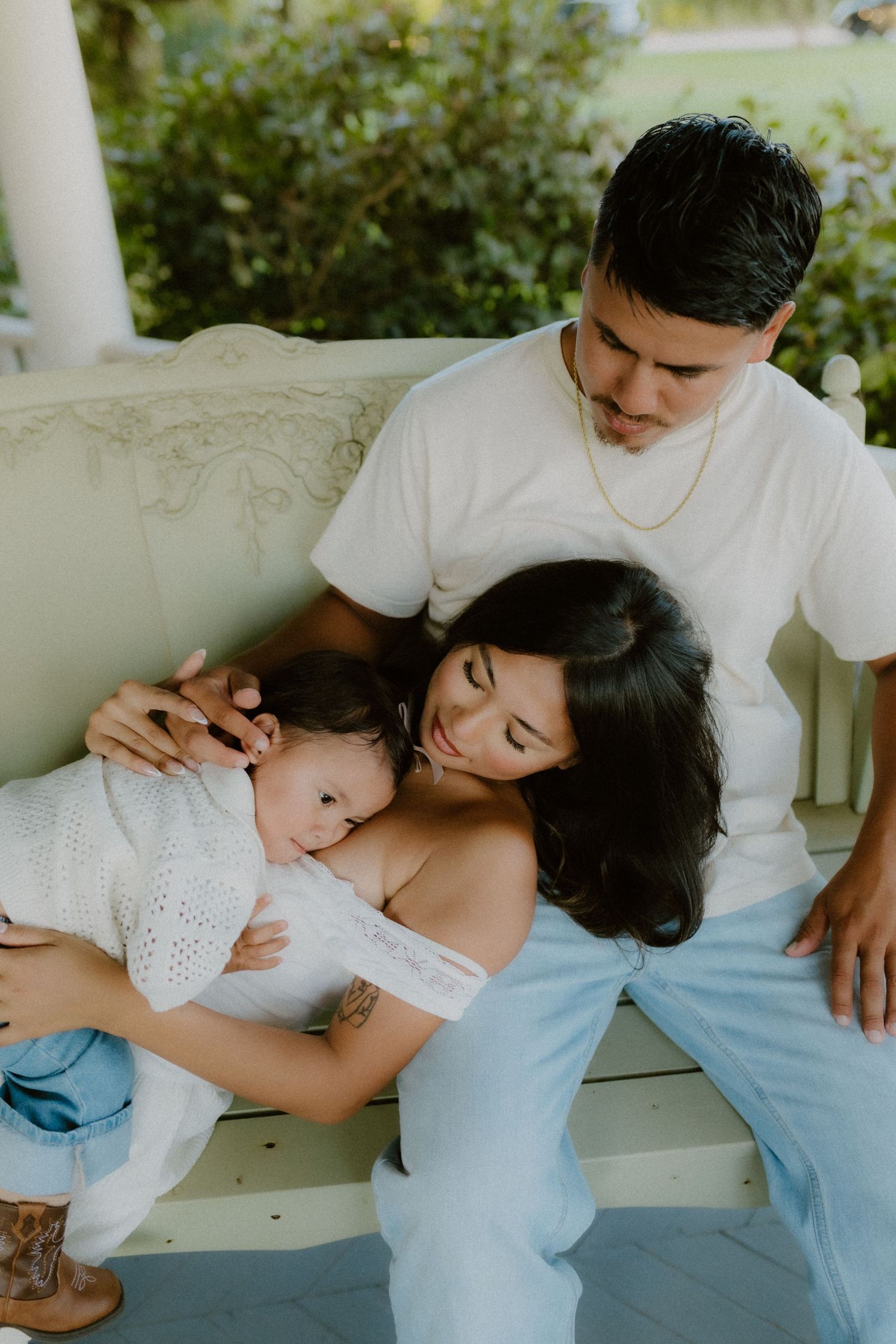 Young parents sit on a porch bench, mother breastfeeding newborn baby while father looks on lovingly.