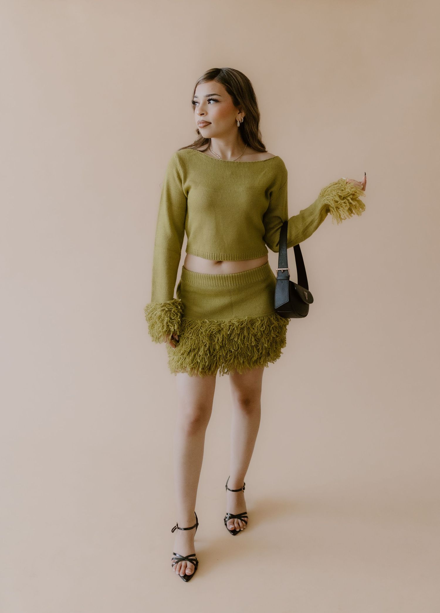 Woman wearing olive green feather-trim crop sweater and mini skirt set with black heels and shoulder bag.
