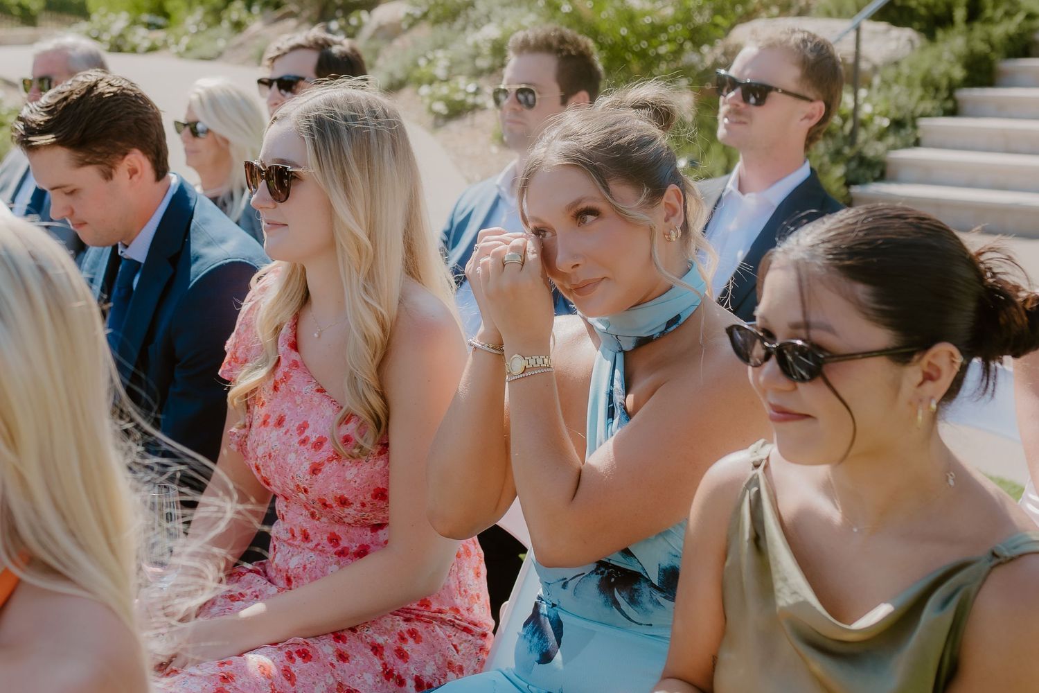 Wedding guests seated outdoors in summer attire, with one woman in blue wiping a tear.