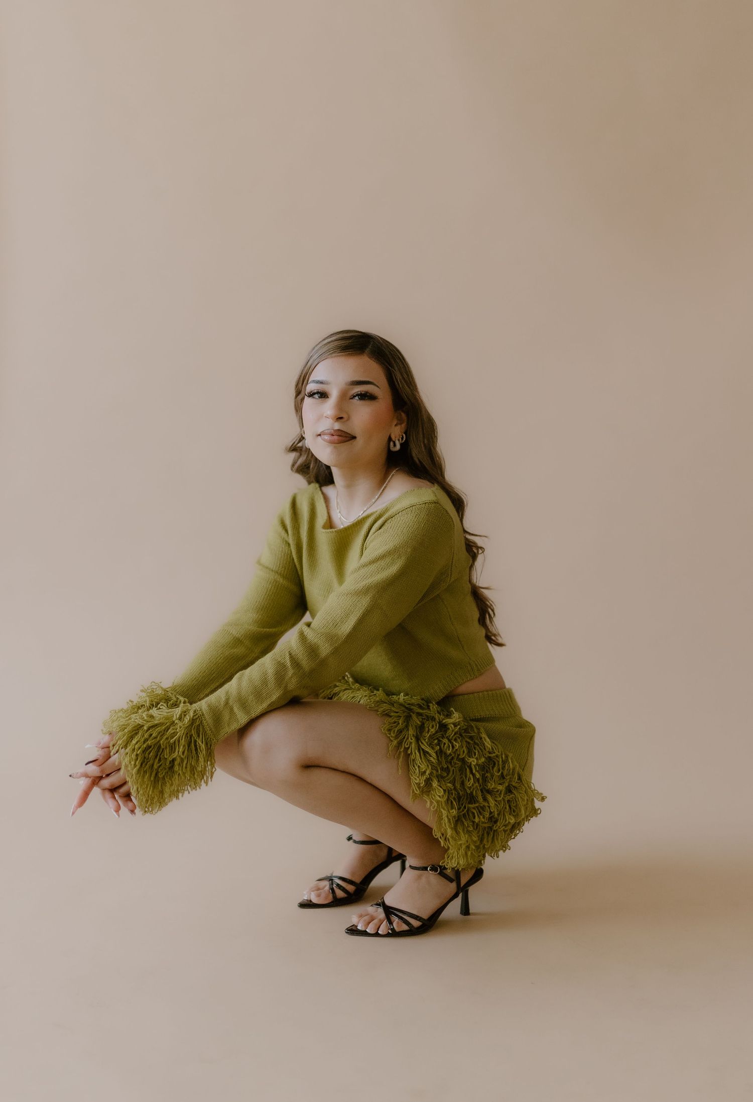 Woman in olive green feathered crop top and mini skirt crouches in black strappy heels on beige backdrop.